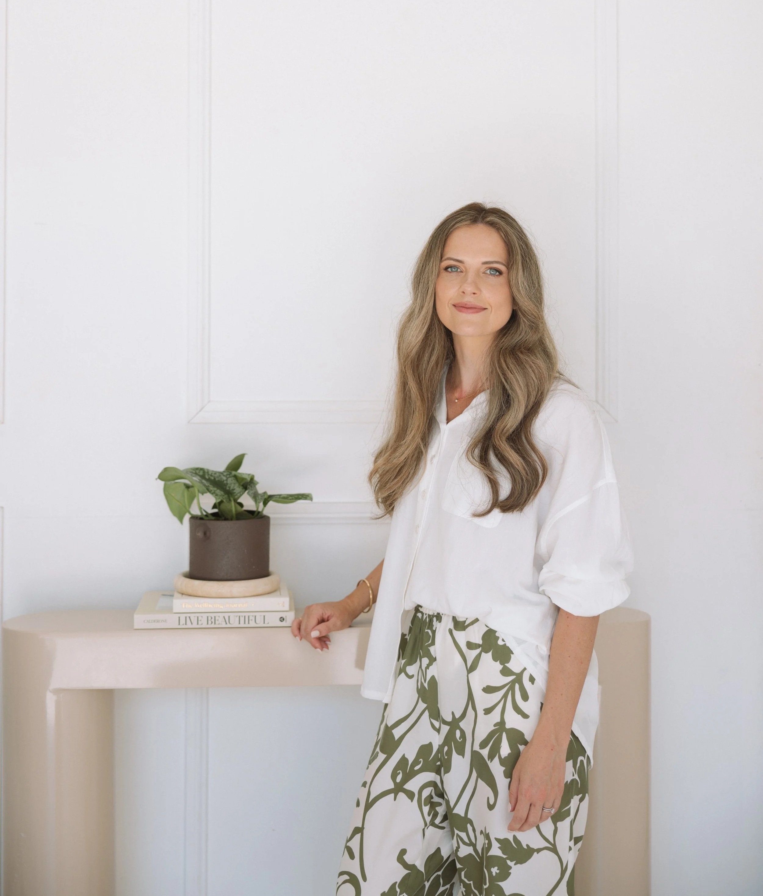A woman with long wavy hair wearing a white button-up shirt and a green and white patterned skirt. She is standing next to a beige table with a potted plant on it, in front of a white wall.
