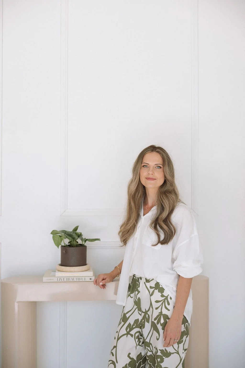 A woman with long wavy hair wearing a white shirt and patterned pants, standing next to a beige table with a potted plant and books, in a white room.