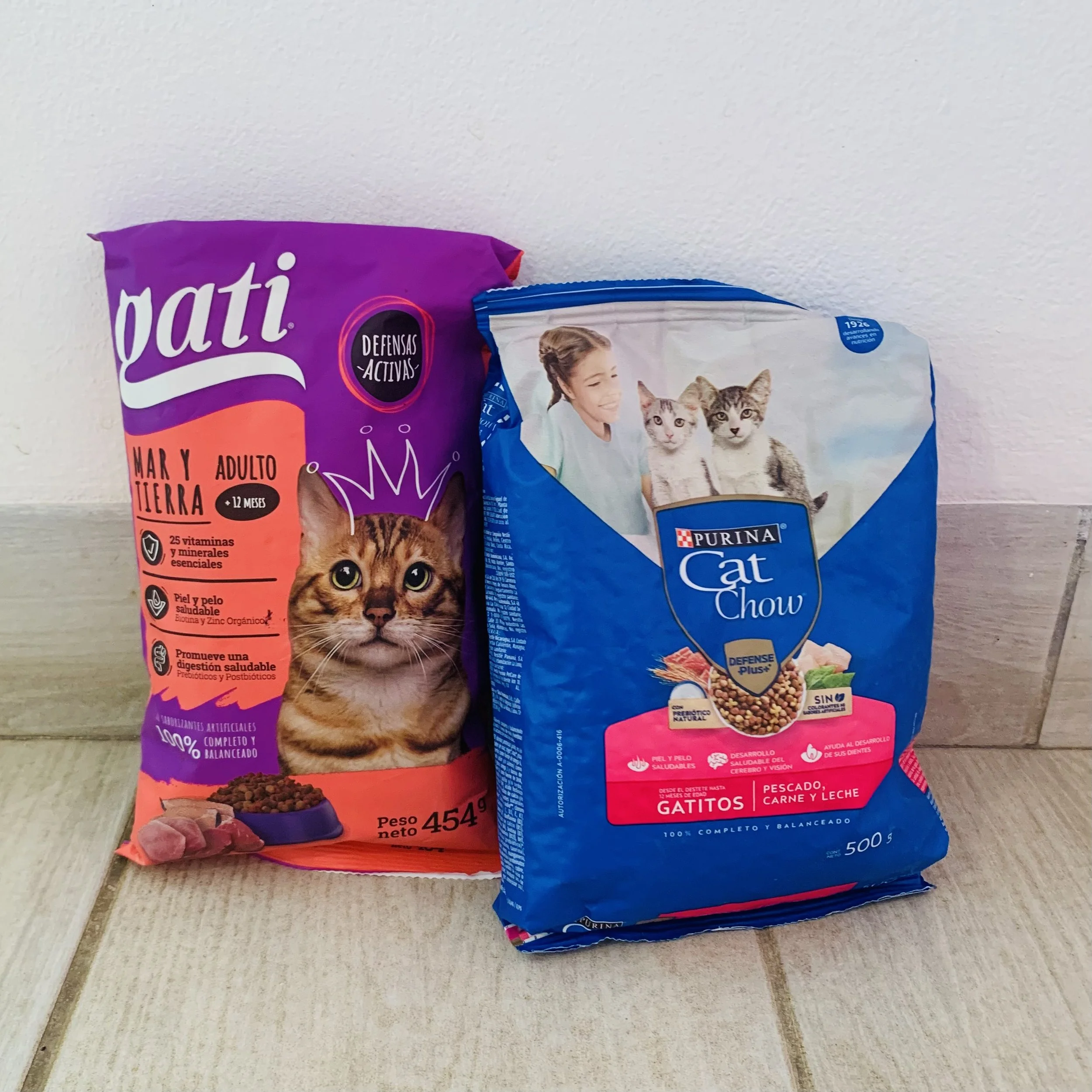 Two bags of pet food, one for cats and one for dogs, placed on a light-colored wooden surface against a plain white wall. The bag on the left features a tabby cat image and has purple and orange coloring, while the bag on the right displays a woman with two kittens and has blue and white coloring.