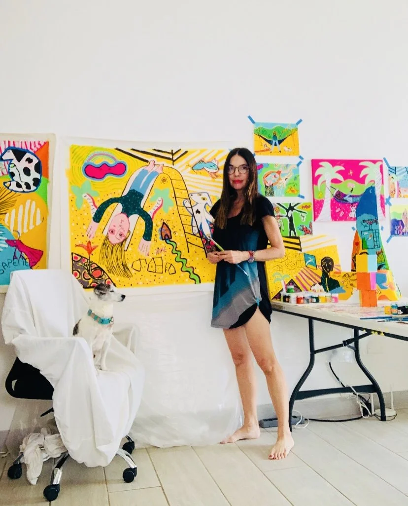 A woman in a black and gray dress holding paintbrushes, standing barefoot next to a dog sitting on a chair covered with a white cloth. The background features colorful, abstract paintings on the wall.