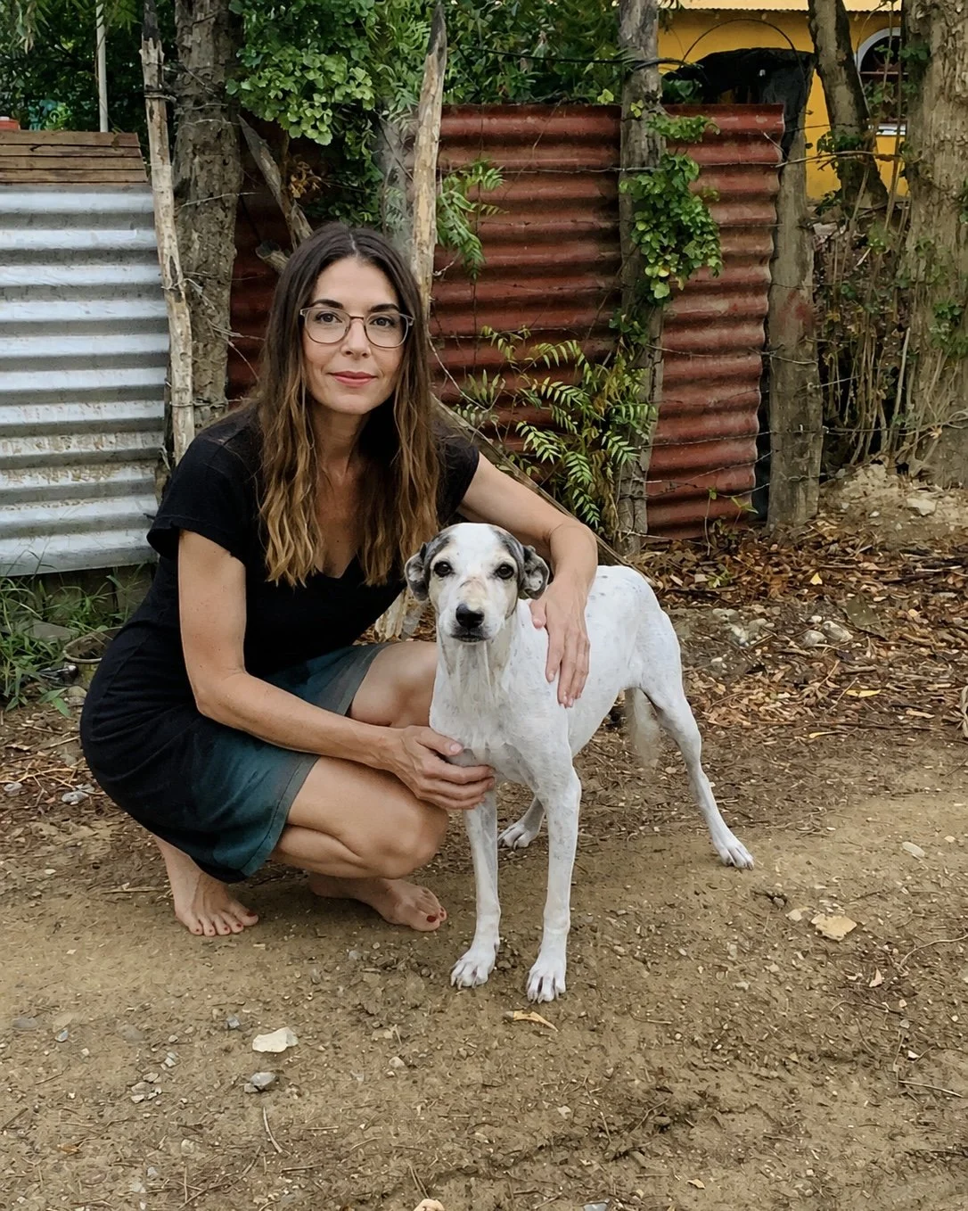 A woman with glasses, wearing a black shirt and shorts, kneeling on the ground outdoors, holding a white dog with black spots. They are in a garden area with dirt ground, trees, and a rustic metal fence in the background.