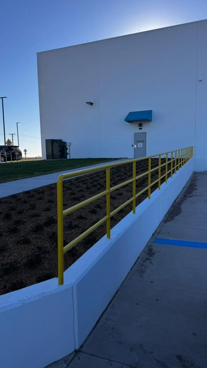 A white building with a blue awning over a door labeled 'W008'. There is a yellow safety railing along a small planting area and a concrete walkway, with a grassy area and a parking lot in the background. The sky is clear and blue.