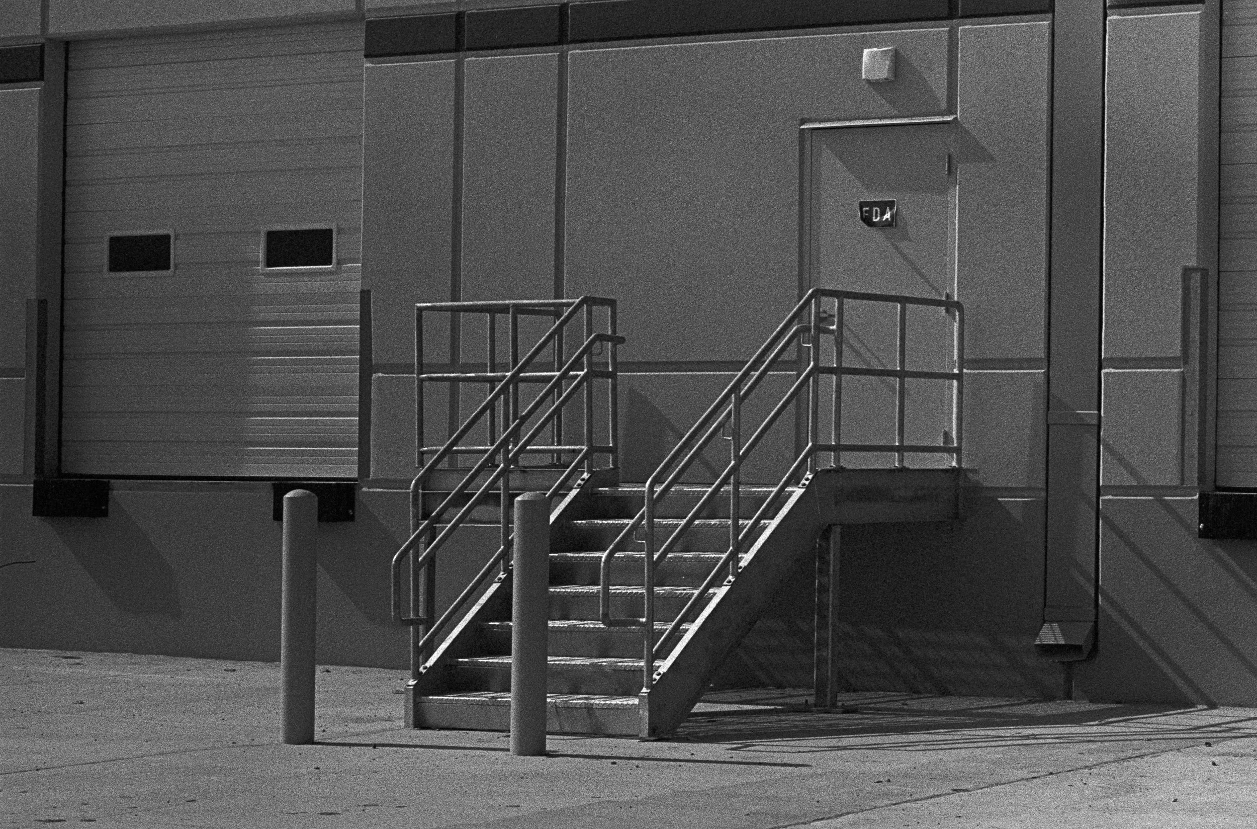 A black and white photo of an industrial building with a set of outdoor stairs leading to a door. There are two small rectangular windows on the left side of the building. The building appears to be made of metal panels.