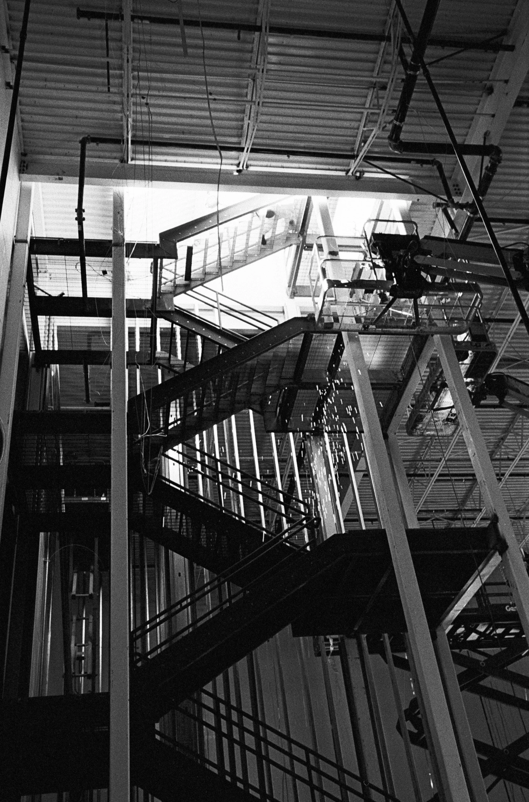 Black and white photo of a multi-level fire escape staircase inside a building with exposed metal pipes and beams.