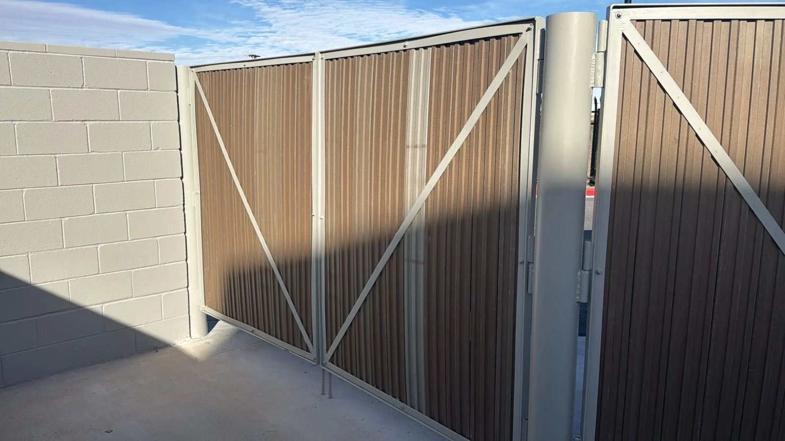 Brown metal gate with a white frame and diagonal support bars, attached to a gray cinder block wall, with a concrete ground and a partly cloudy blue sky in the background.