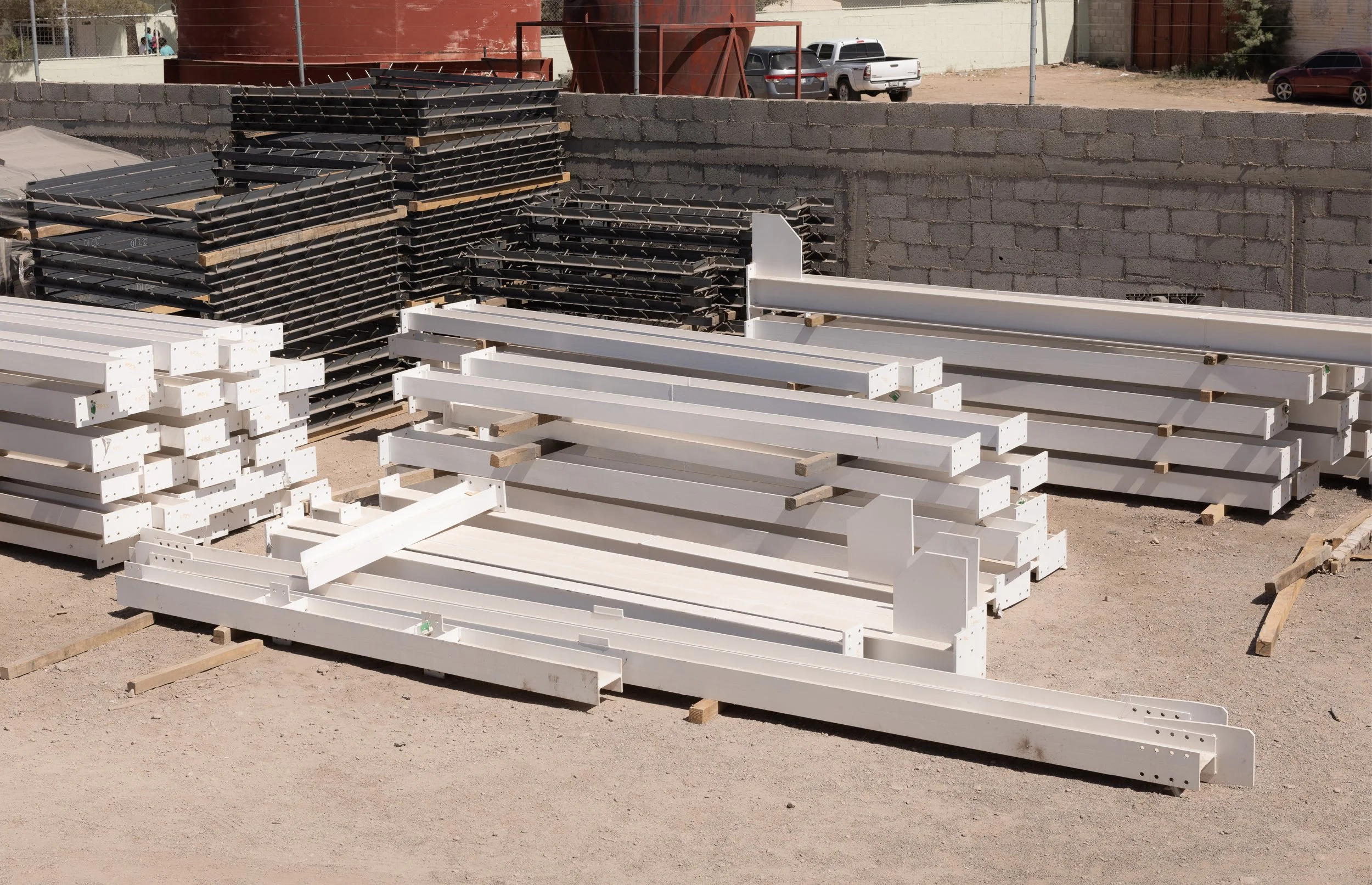 Stacks of construction materials including white metal beams and black metal panels stored in an outdoor construction site with a brick wall and parked cars in the background.