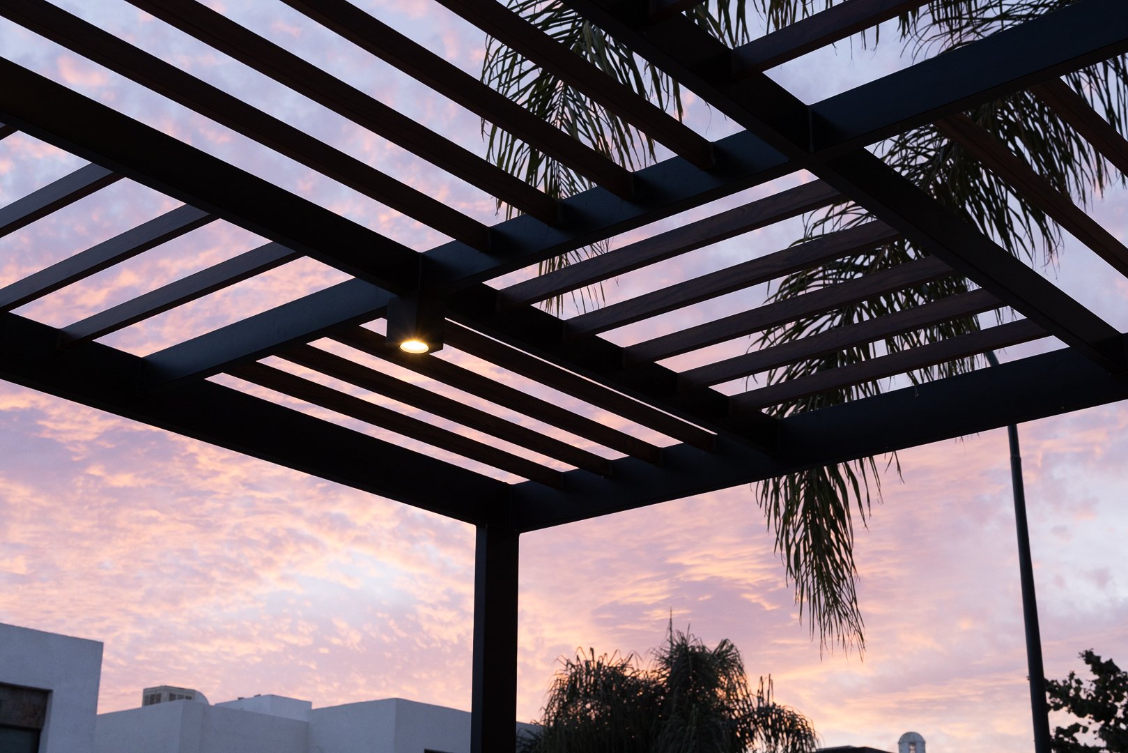 A black metal pergola with horizontal slats against a pink and purple evening sky, with leaves and trees in the background.