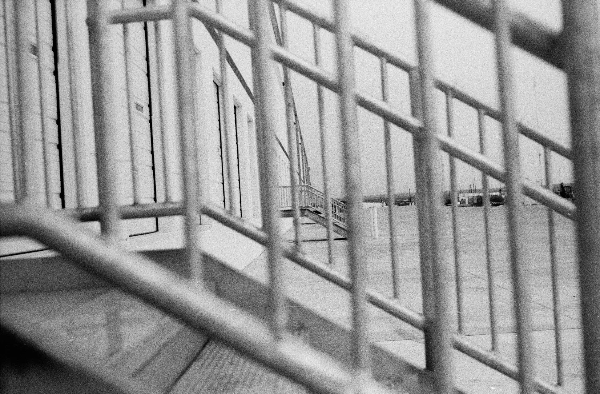 Black and white photo viewed through metal bars of a prison or detention cell, with buildings or structures in the background