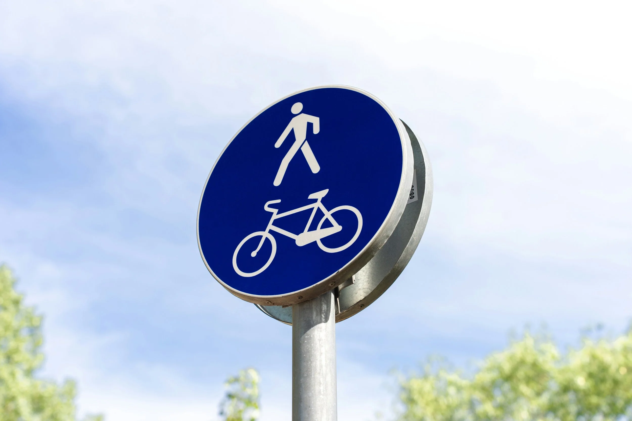 Blue circular sign with white symbols of a pedestrian and a bicycle, indicating a shared path for walking and biking, against a partly cloudy sky with blurred greenery at the bottom.