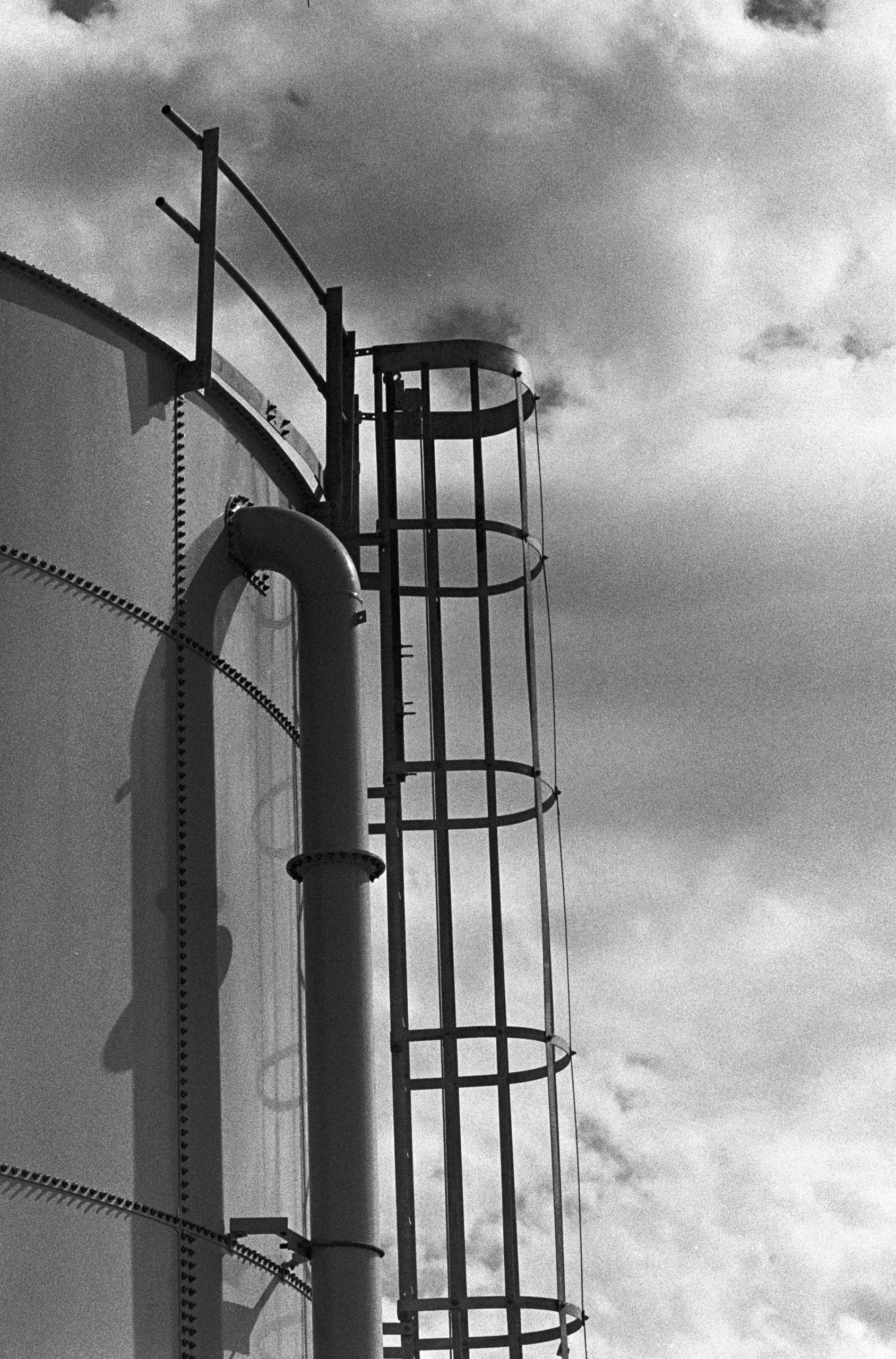 Black and white photo of an industrial structure with cylindrical tanks and a ladder or spiral staircase on the right, against a clouded sky.