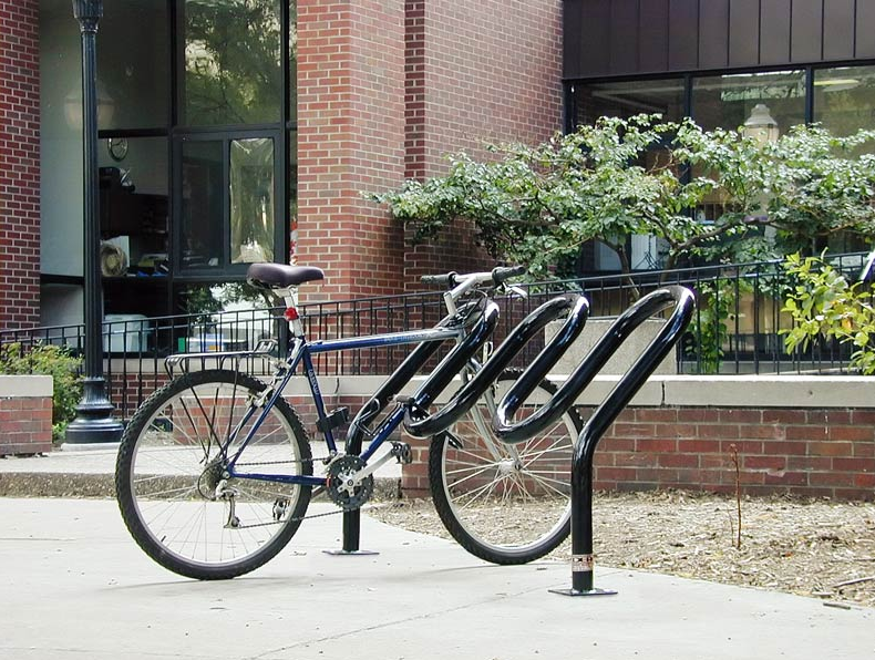A blue bicycle locked to a black bike rack outside a brick building with windows and green plants.