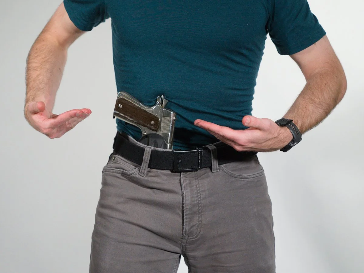 A person wearing a teal T-shirt and gray pants with a gun that is too large for their frame tucked into their waistband, with their hands gesturing around the gun.