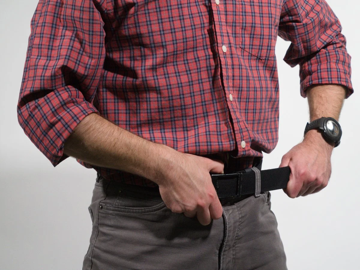 A man wearing a red plaid shirt and gray pants adjusting his black belt. He is also wearing a watch on his left wrist.