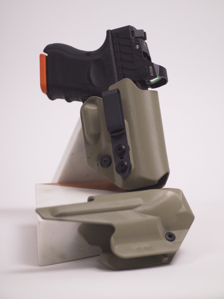 A black handgun in a foliage green Mockingbird Defense holster, with a reversed holster resting below, mounted on a beige display stand on a white background.