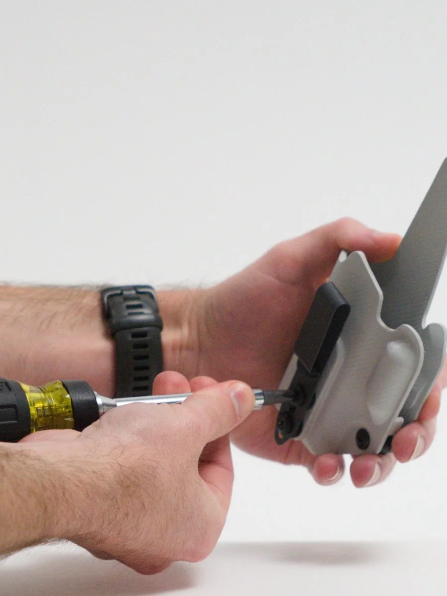 Person tightening screws on a black belt clip onto a gray Mockingbird Holsters holster using a screwdriver held between the thumb and fingers, against a plain white background.