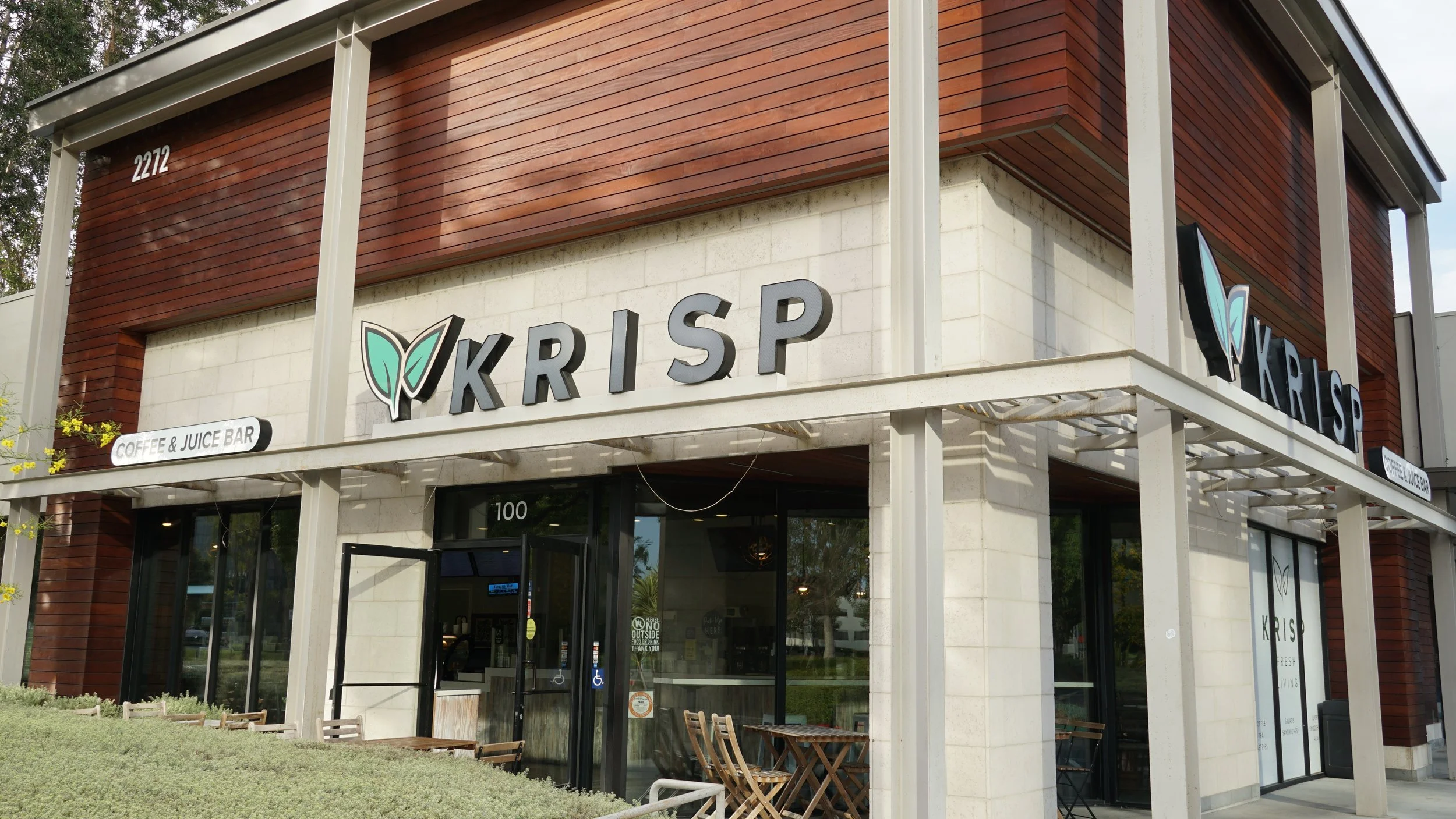 Exterior of Krisp coffee and juice bar with a modern design, wooden and stone facade, outdoor seating, and signage displaying the name Krisp and services.