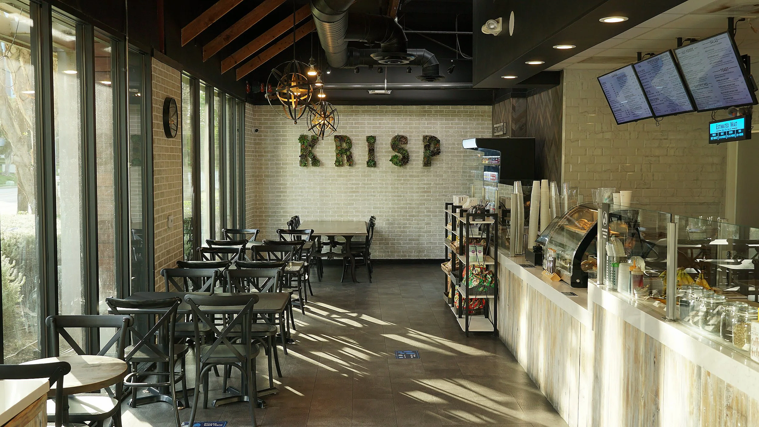 Inside of a modern coffee shop with black chairs and tables, large windows, a counter area with a food display, multiple digital menu screens, and a decorative wall with the word 'KRISP' spelled out with greenery.