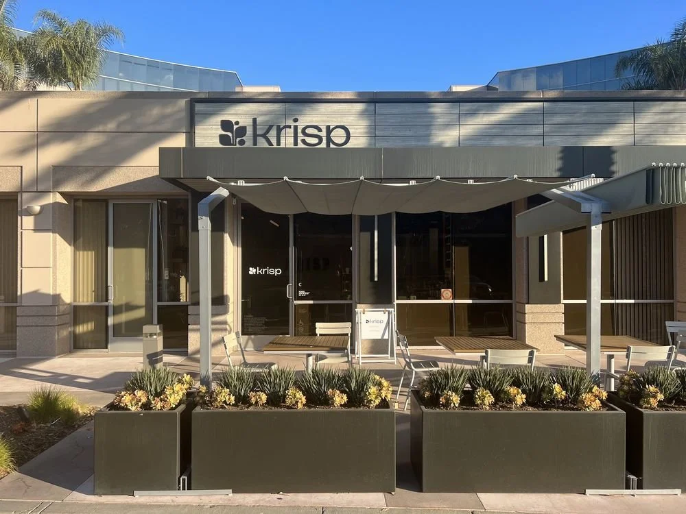 Exterior of a Krisp office with a modern facade, outdoor seating, and planters with decorative plants, under a clear blue sky.