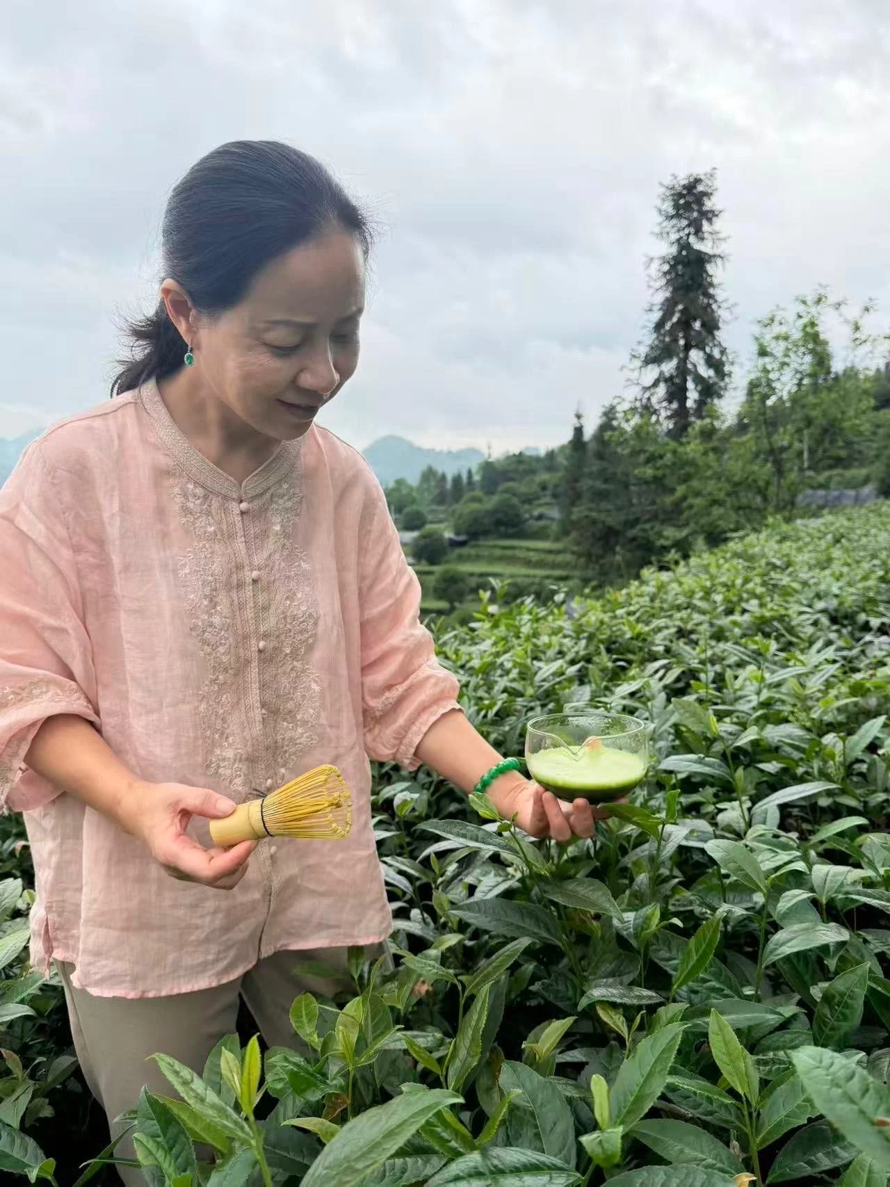Woman in a pink blouse holding a tea whisk and a cup of matcha tea in a green tea plantation.