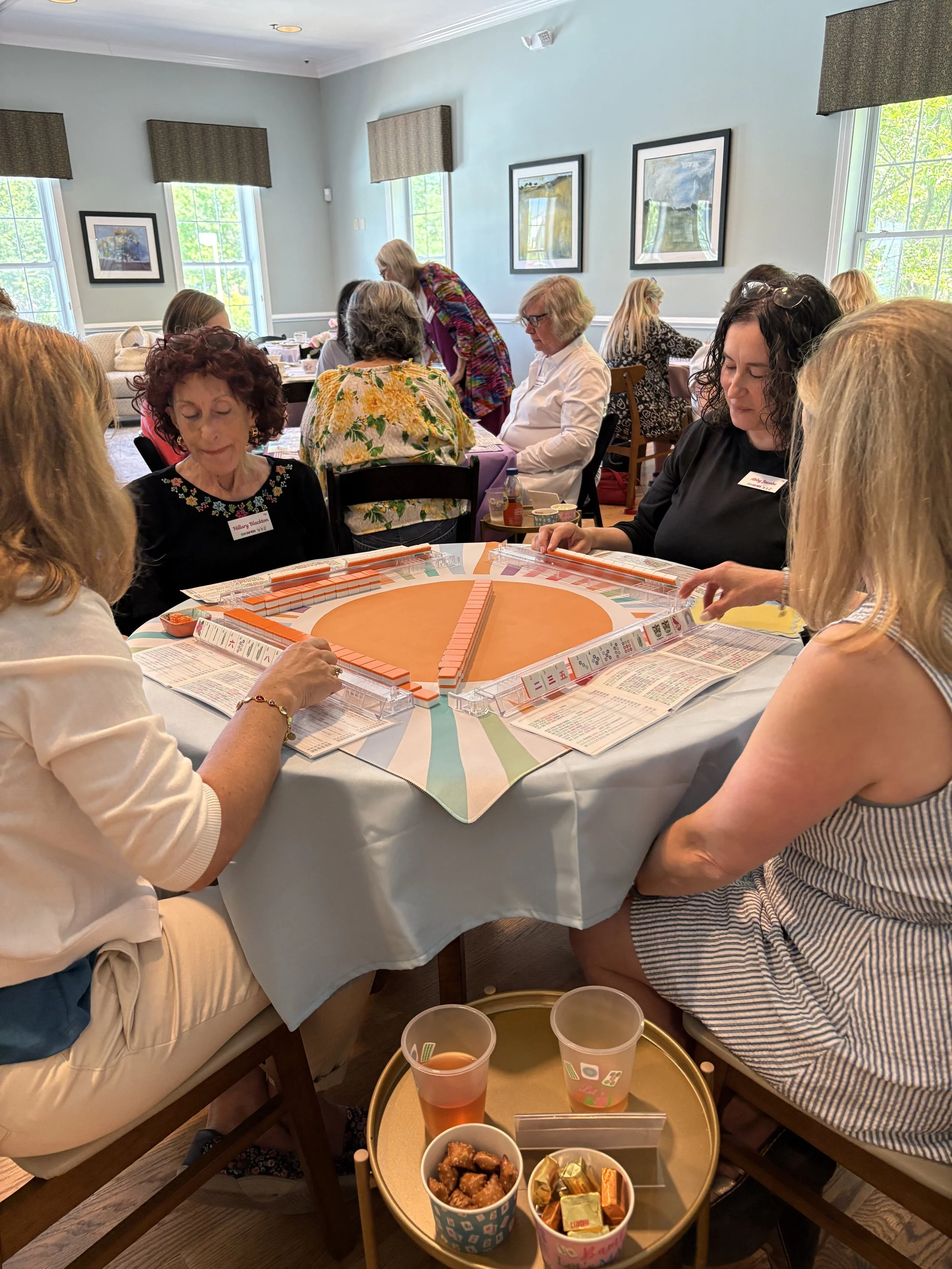 A group of women playing a game around a circular table during a social event. The table has snacks and drinks, and women are engaged and smiling.