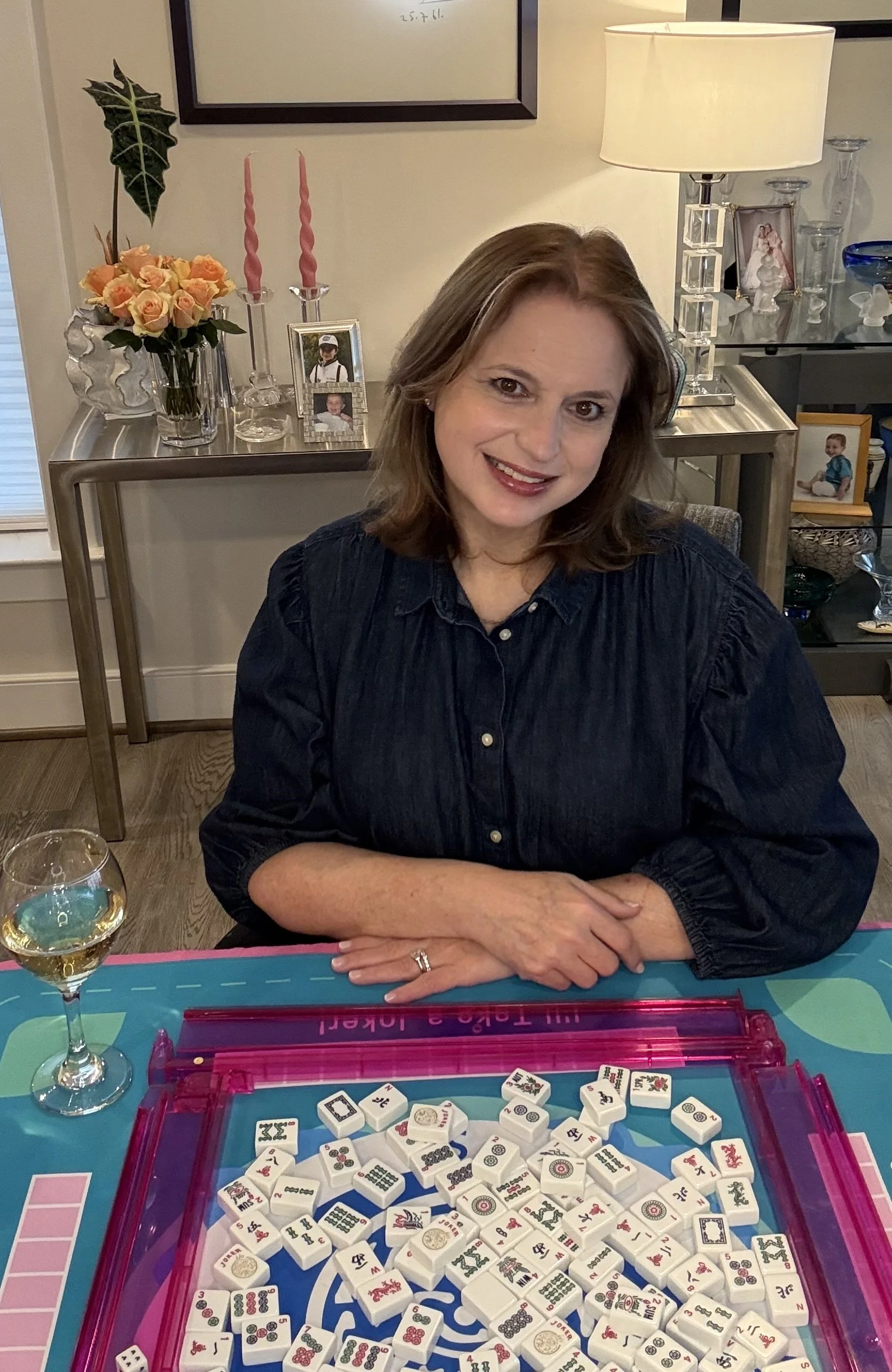 A woman sitting at a table with a glass of white wine, playing Mahjong with tiles on a colorful mat in front of her, in a cozy, well-decorated room with family photos, a lamp, and floral arrangements in the background.
