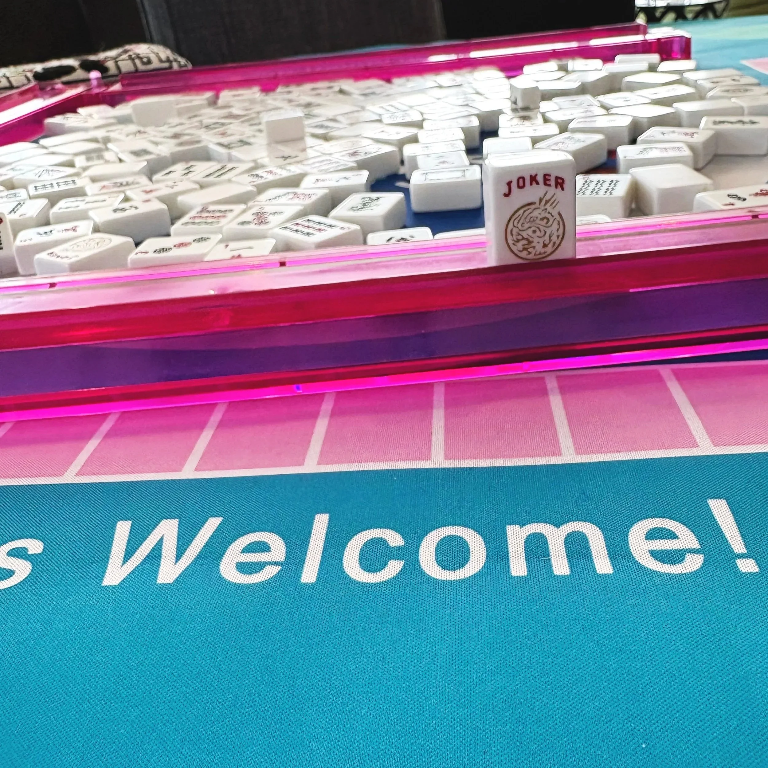Close-up of a Mahjong game with white tiles inside a pink plastic case, a joker tile with a dragon design in the front, and a blue game mat with the word 'Welcome!' visible.