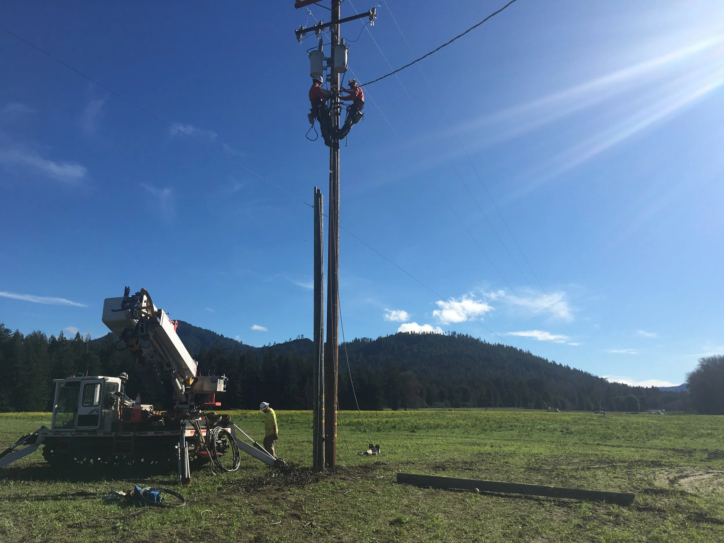 IBEW linemen working on a utility pole in a green mountain valley, with a bucket truck and crew on the ground below a clear blue sky.
