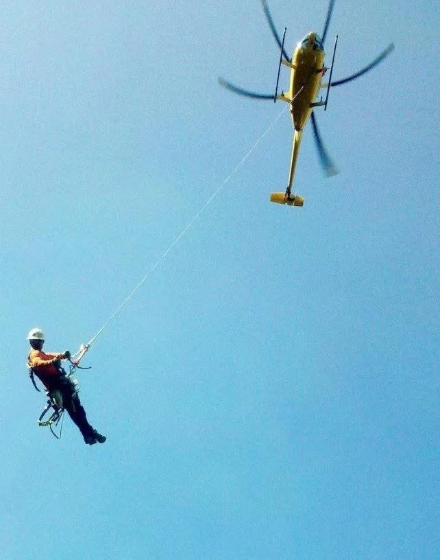 IBEW outside line worker being hoisted into the air by a helicopter using a long cable against a clear blue sky.