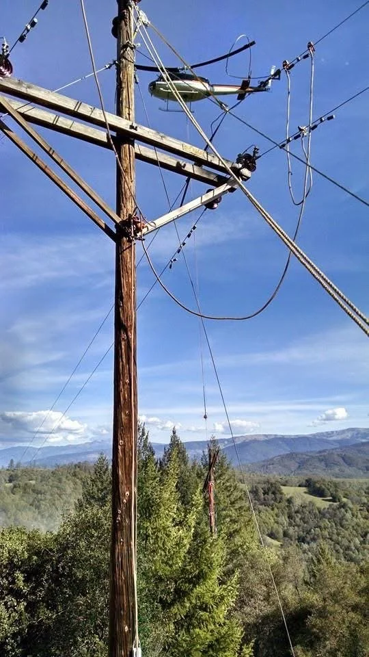 A wooden utility pole with electrical wires and a transformer mounted on top, set against a backdrop of green trees and mountains under a partly cloudy sky and helicopter in the background.
