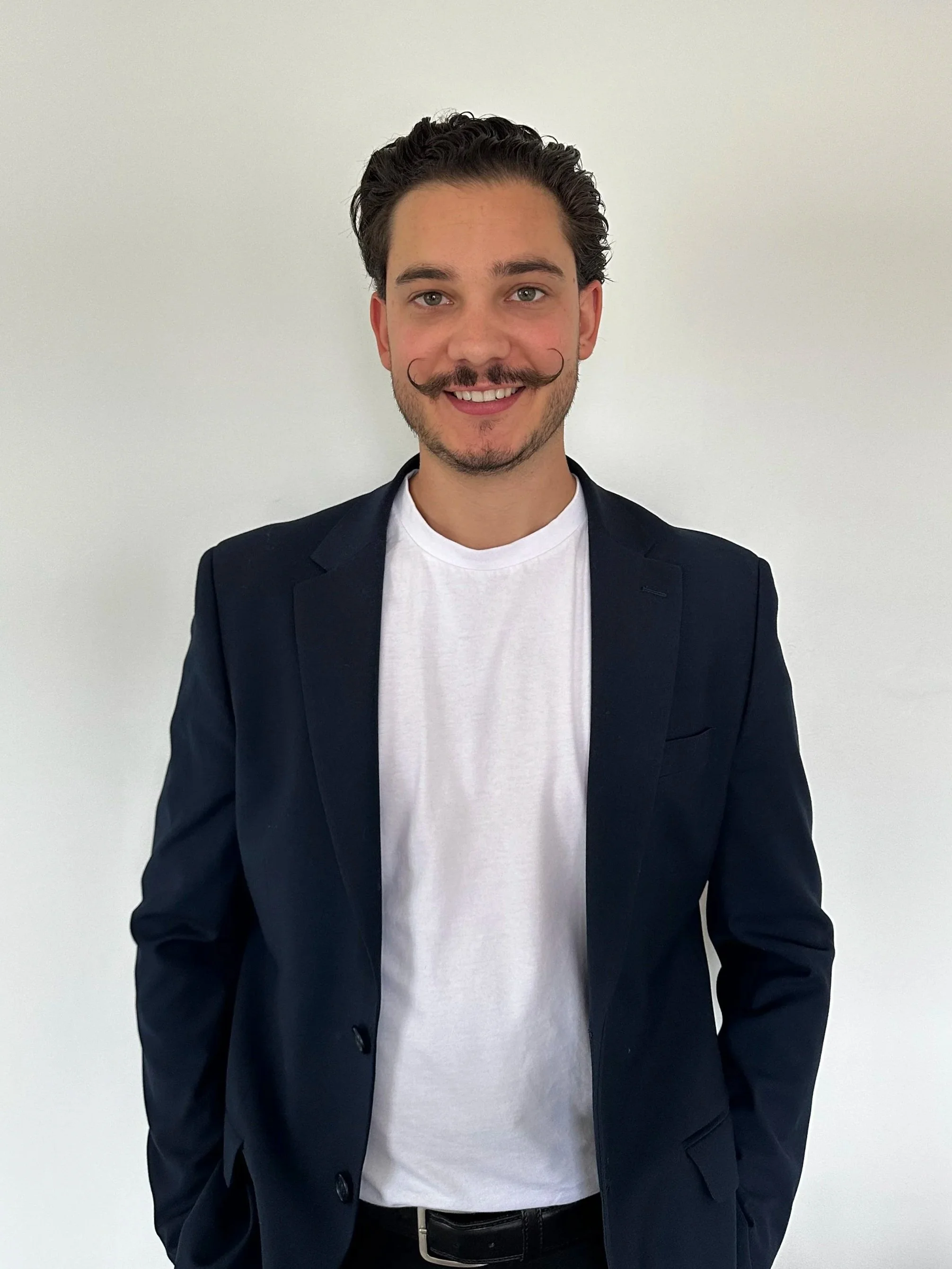 A young man with dark, curly hair and a styled mustache. He is smiling and dressed in a white t-shirt and a navy blue blazer, standing against a plain light-colored background.