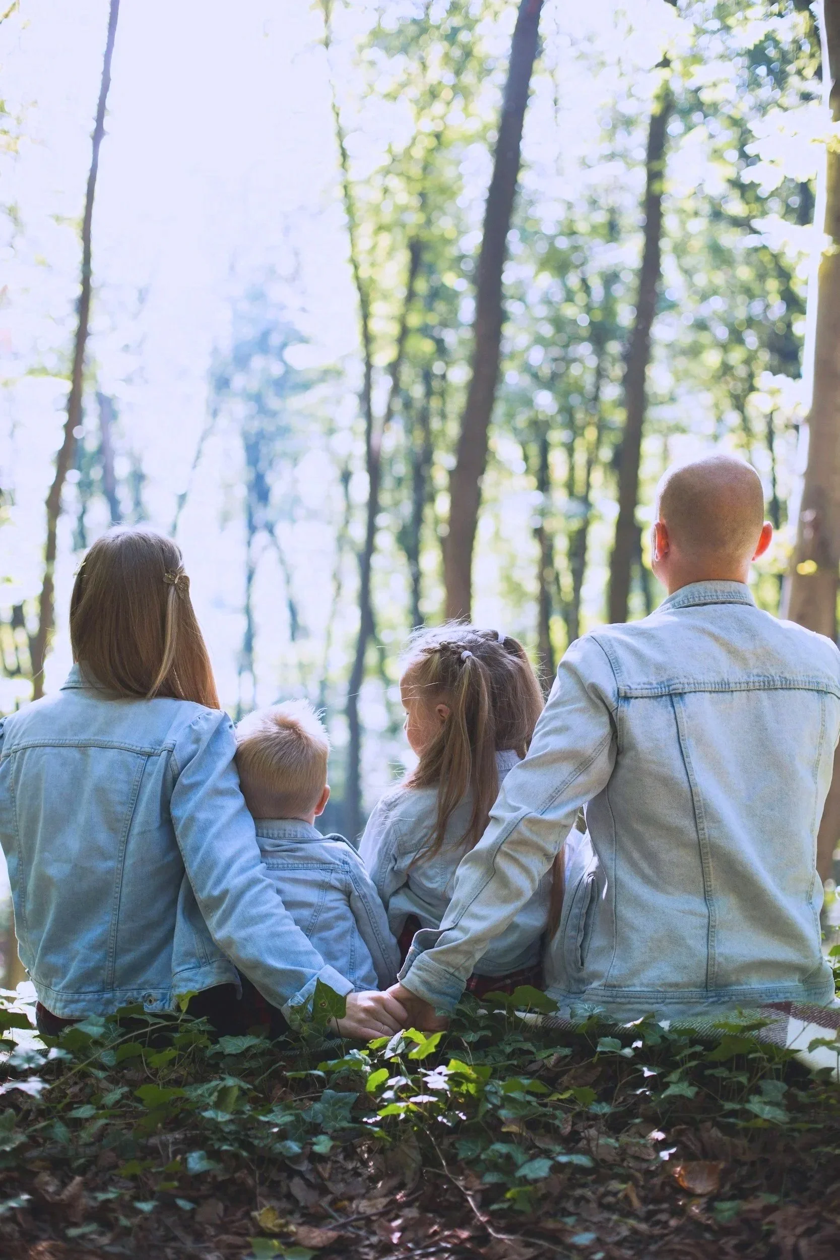 A group of people, including children and adults, standing in a circle holding hands in a forest with tall trees and green foliage, participating in an outdoor activity.