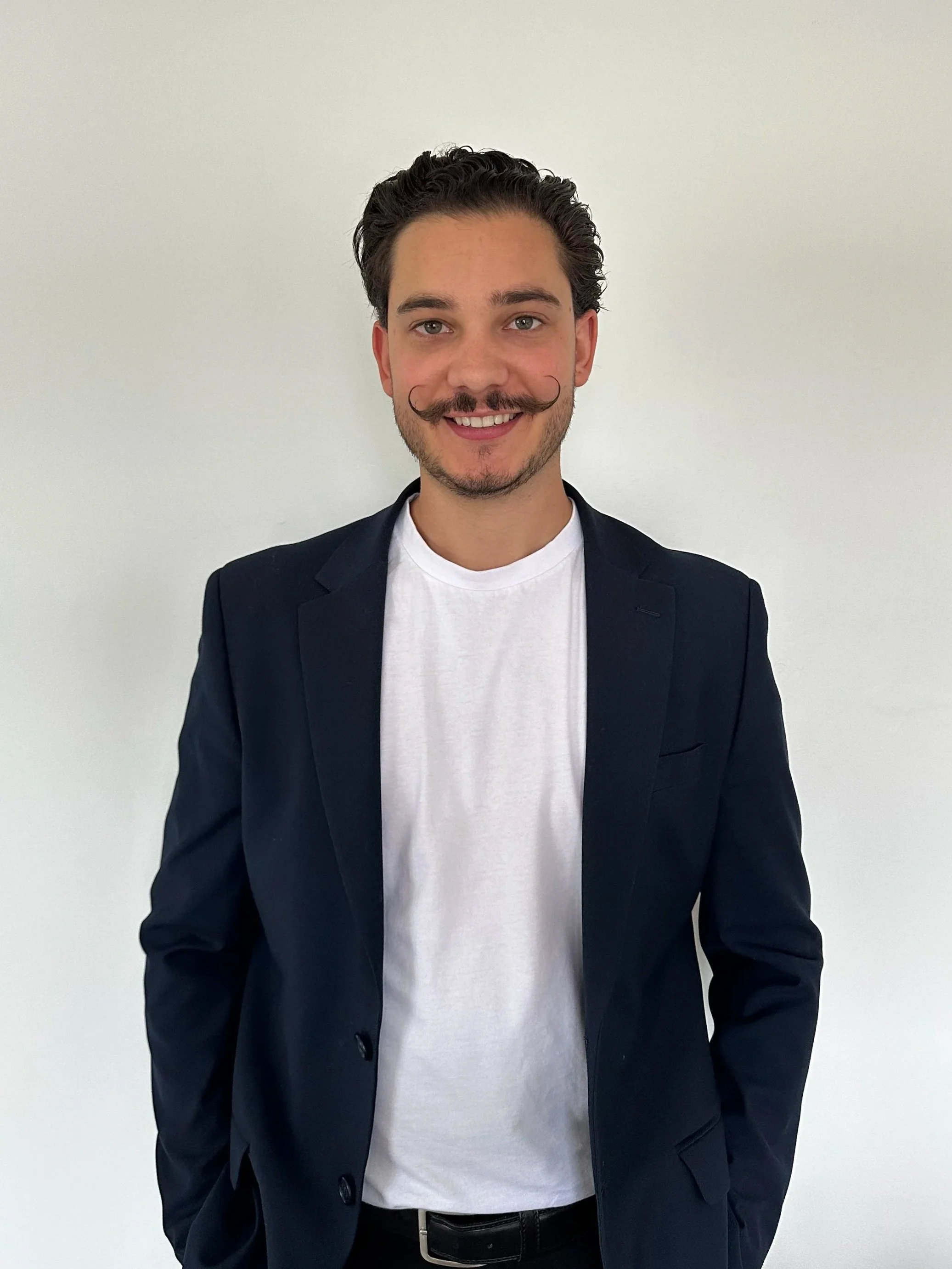 A smiling young man with a mustache, dressed in a dark blazer and white T-shirt, standing against a plain white wall.