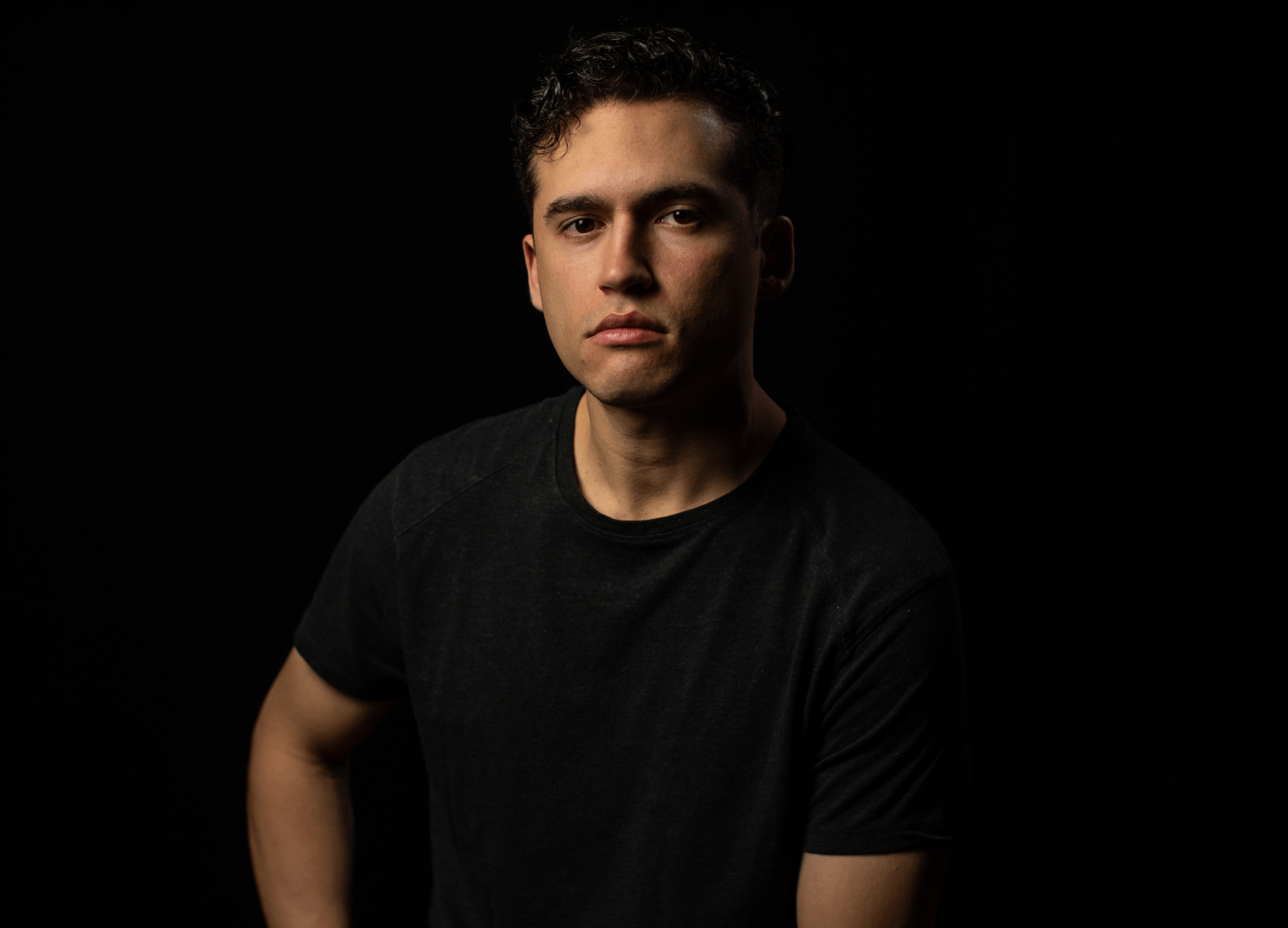 A young man with dark hair and a serious expression, wearing a black t-shirt, posing against a black background.