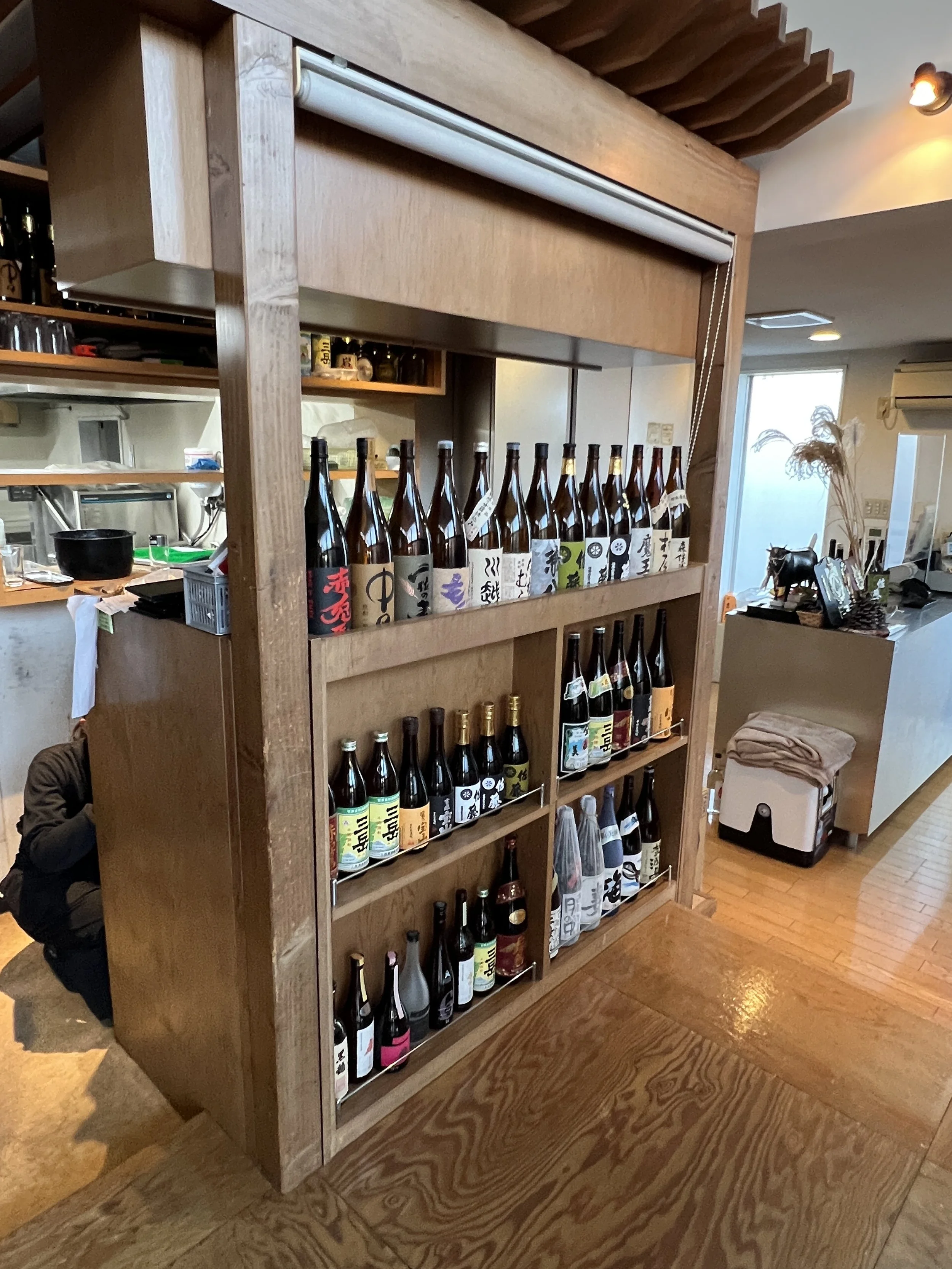 木製の棚に並べられたさまざまな種類の日本酒の bottles。店内の一部の風景も見える。
