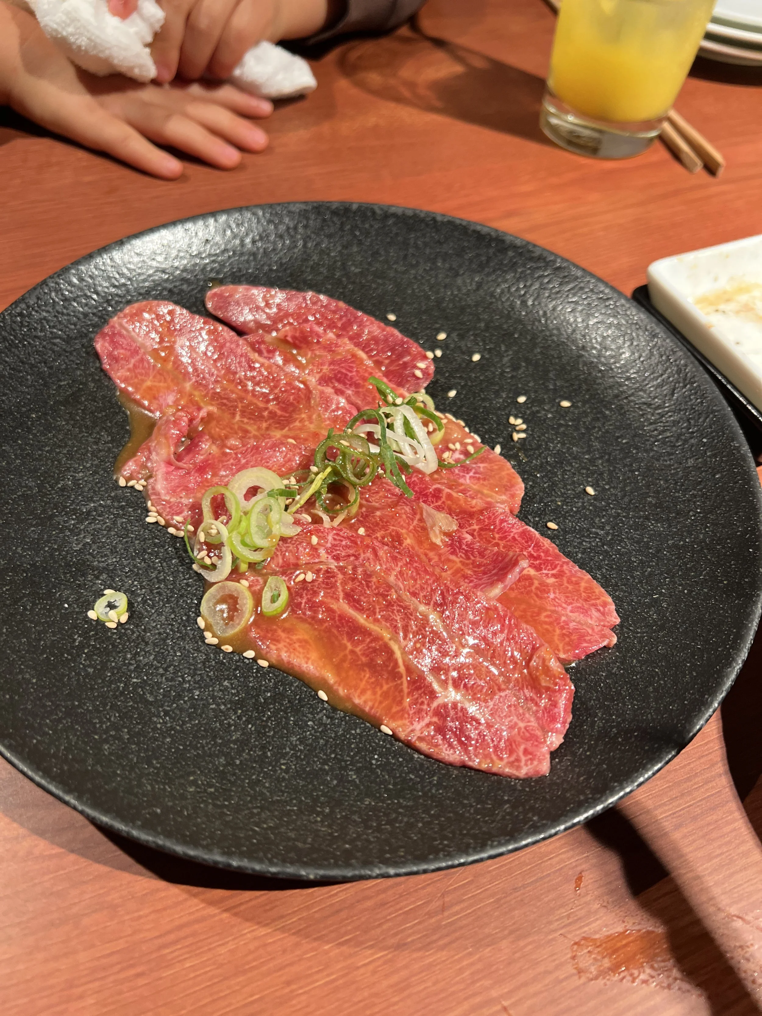 焼肉のための薄切り牛肉とネギの盛り付けがされた黒いプレート。背景に飲み物のグラスと手、その他の皿が見える。