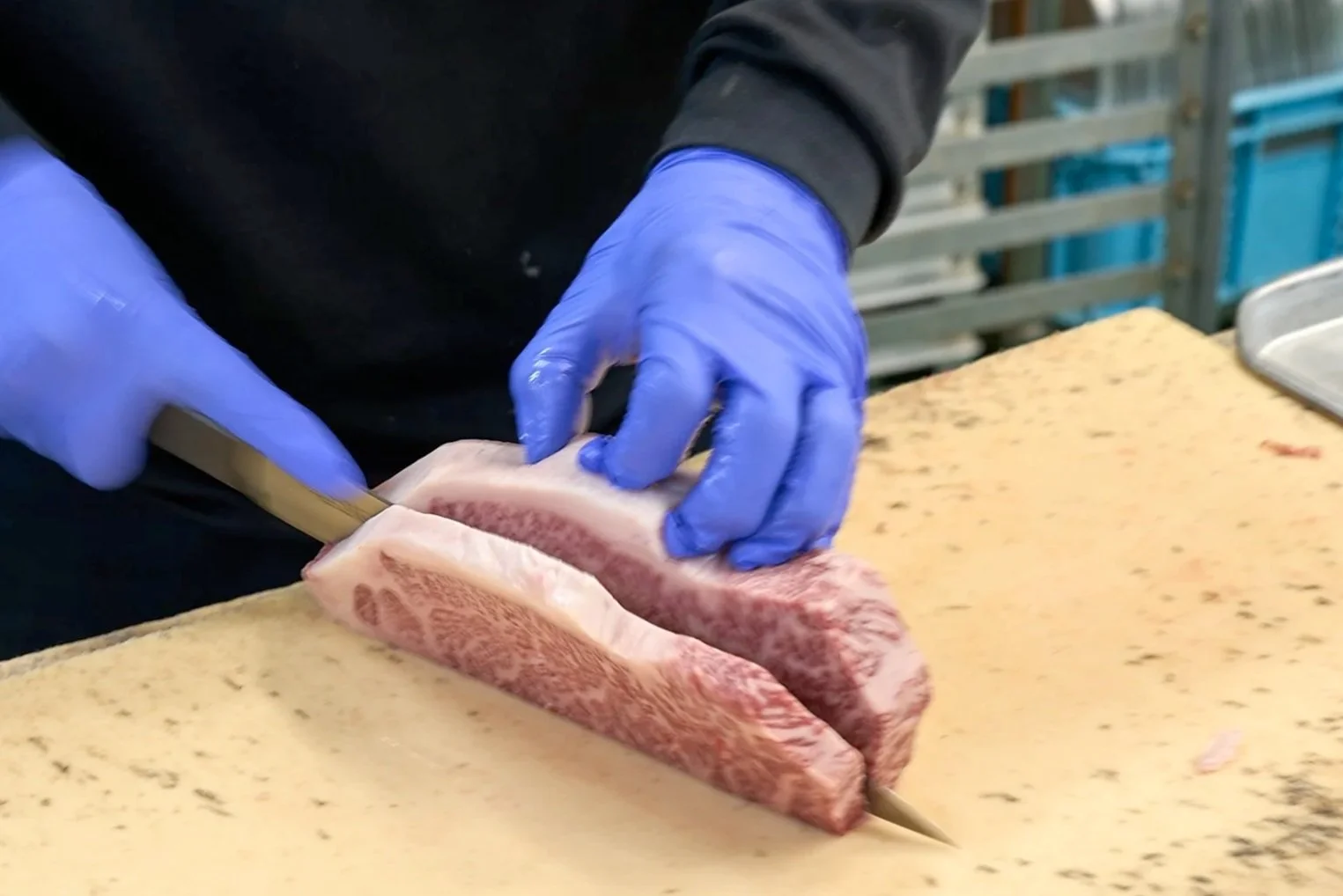 青い手袋をした人物が、肉の塊を包丁でさばいている様子。