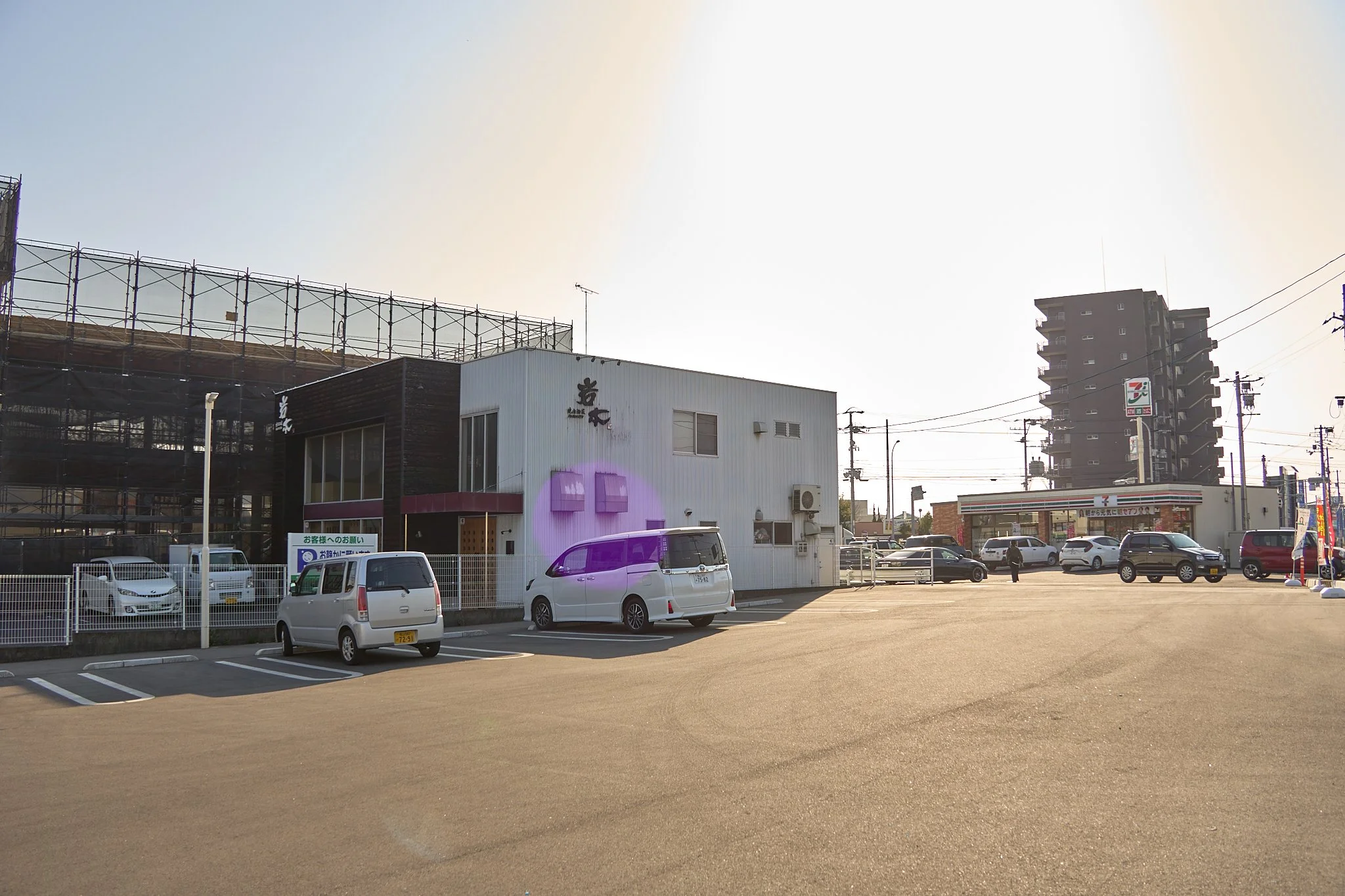 広い駐車場と商業施設、セブンイレブンのコンビニエンスストア、マンションの高層建築物が見える。