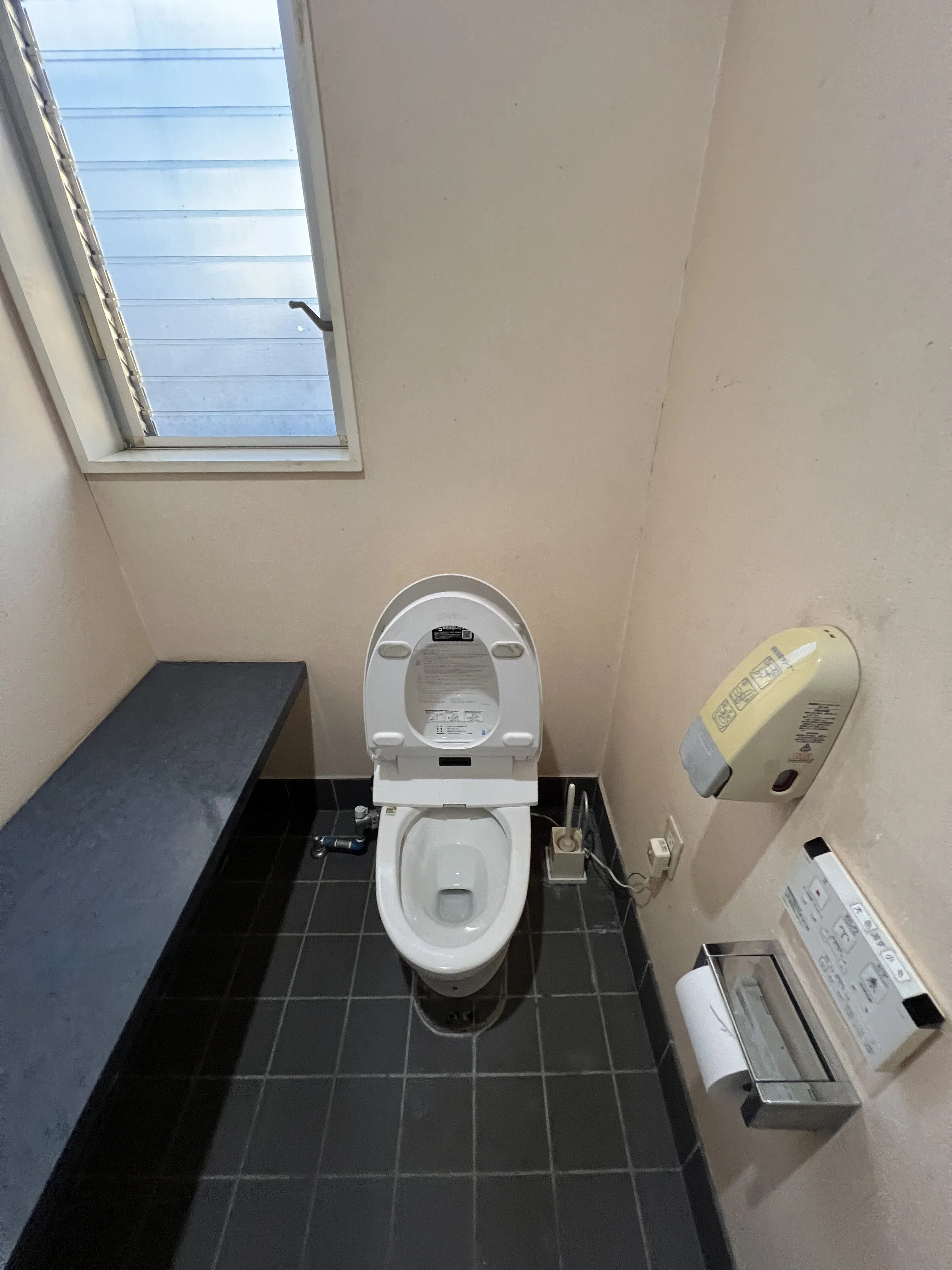 小さなトイレルームの中にあり、壁に設置された紙巻きトイレットペーパーホルダー、洗浄便座の便器、窓、電気プラグとホース、紙巻器、壁掛けのウォシュレット用装置が見えるシンプルなトイレの写真です。