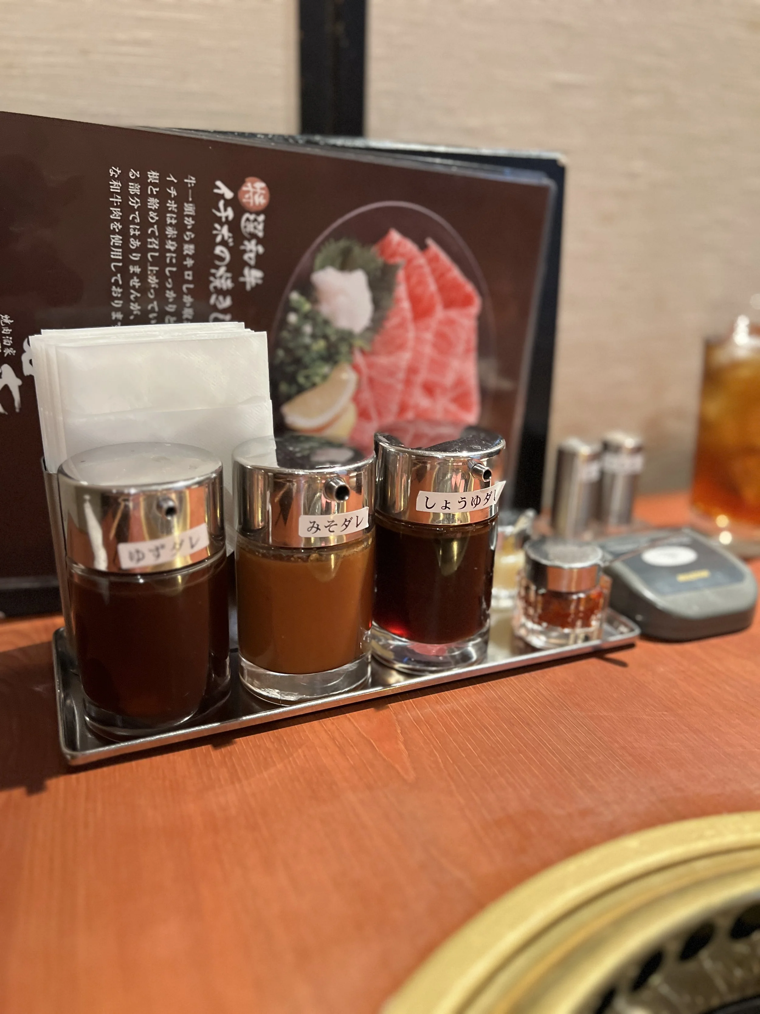 テーブルの上に醤油、みそダレ、つゆダレの容器と白い紙ナプキンが置かれた様子と、背後にメニューと焼肉の写真がある。