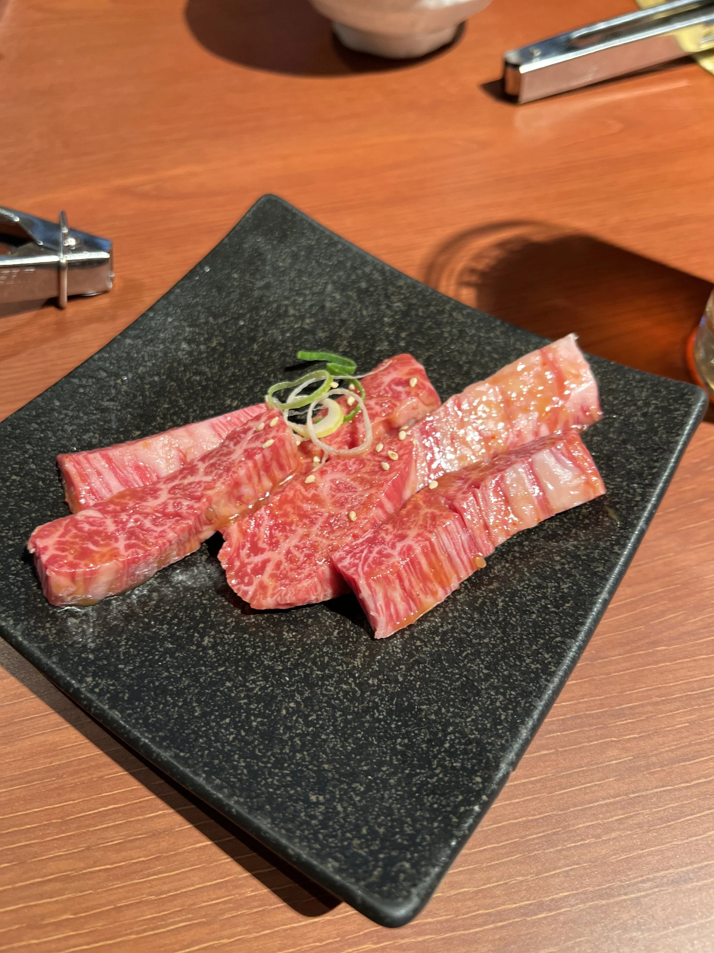 焼肉用の薄切り牛肉が黒い角皿に盛られている。上にねぎとゴマがトッピングされている。