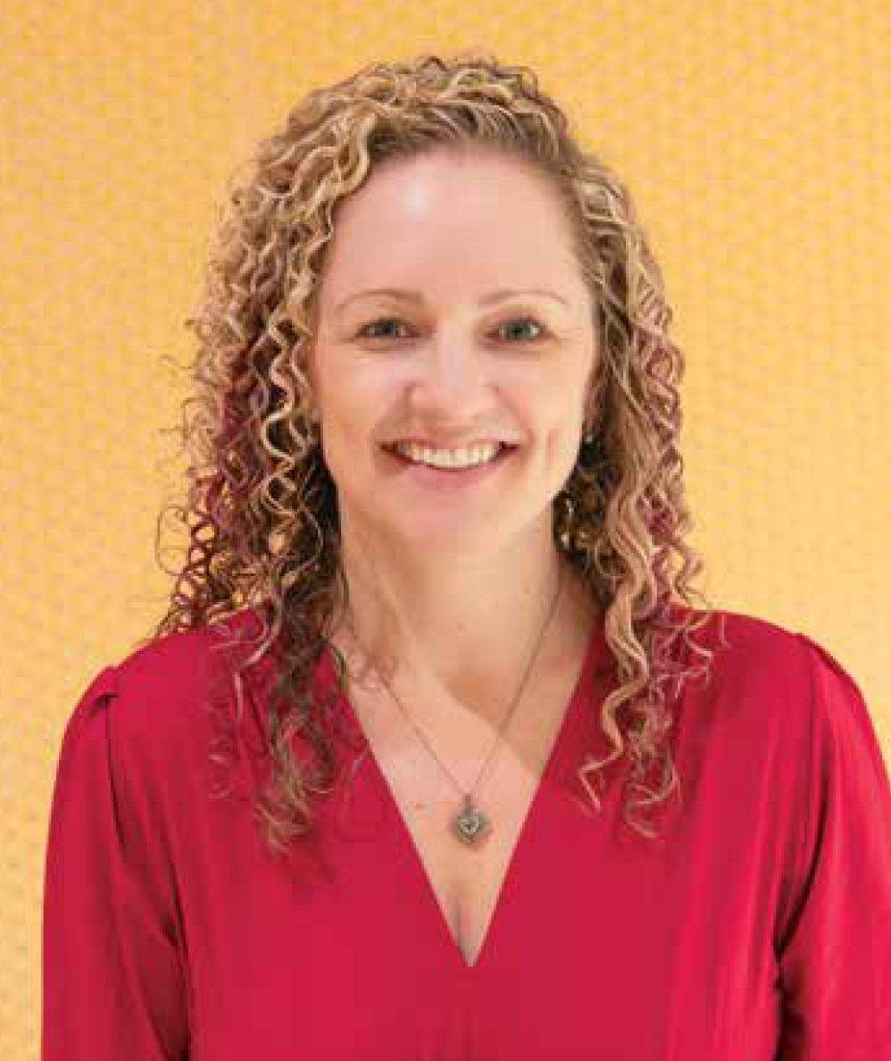 A woman with long curly blonde hair wearing a red top and a necklace, smiling against a yellow background.