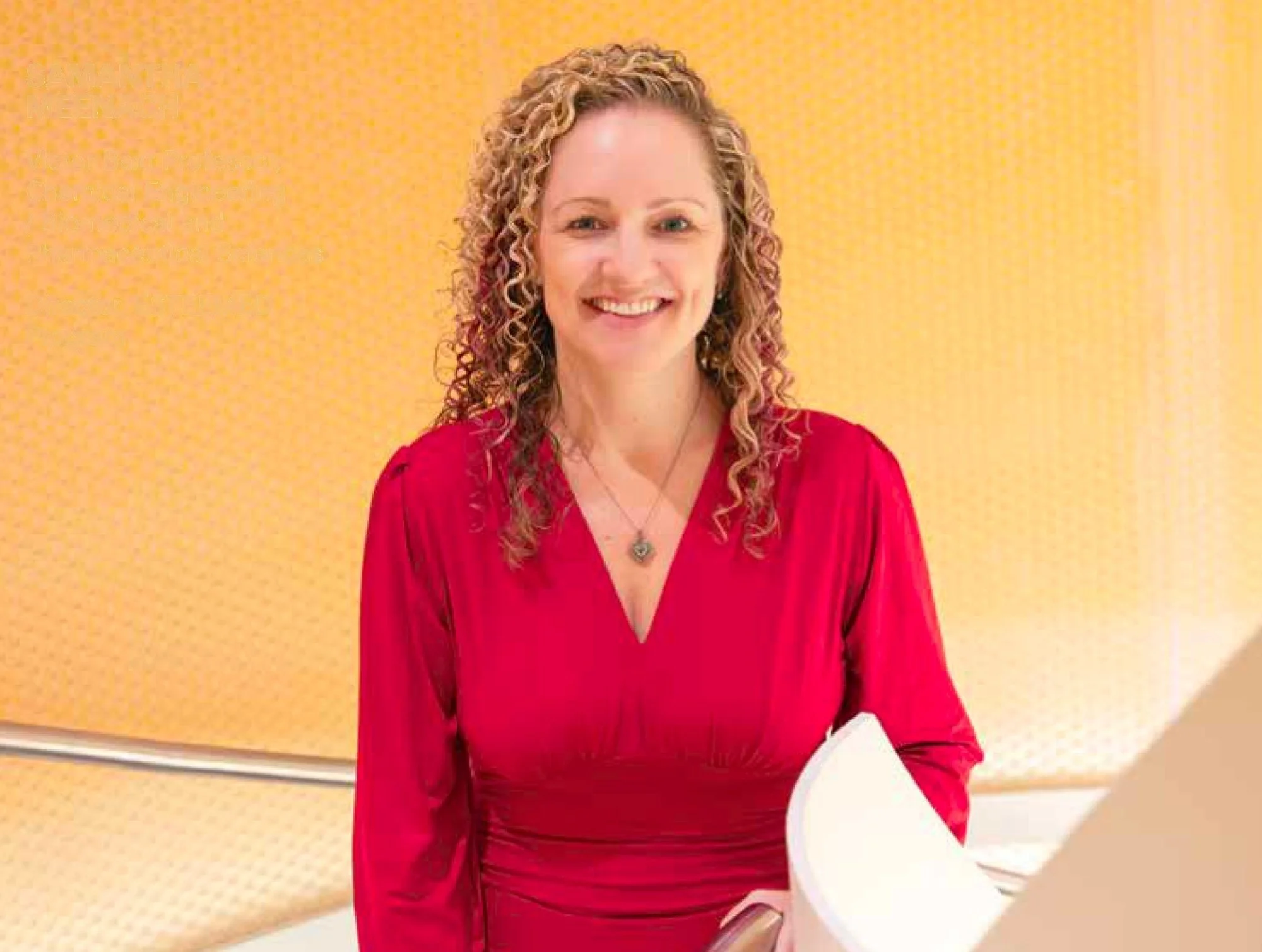 A woman with curly blonde hair wearing a red dress and a silver necklace, smiling, standing in front of a yellow wall with a microphone in front of her.