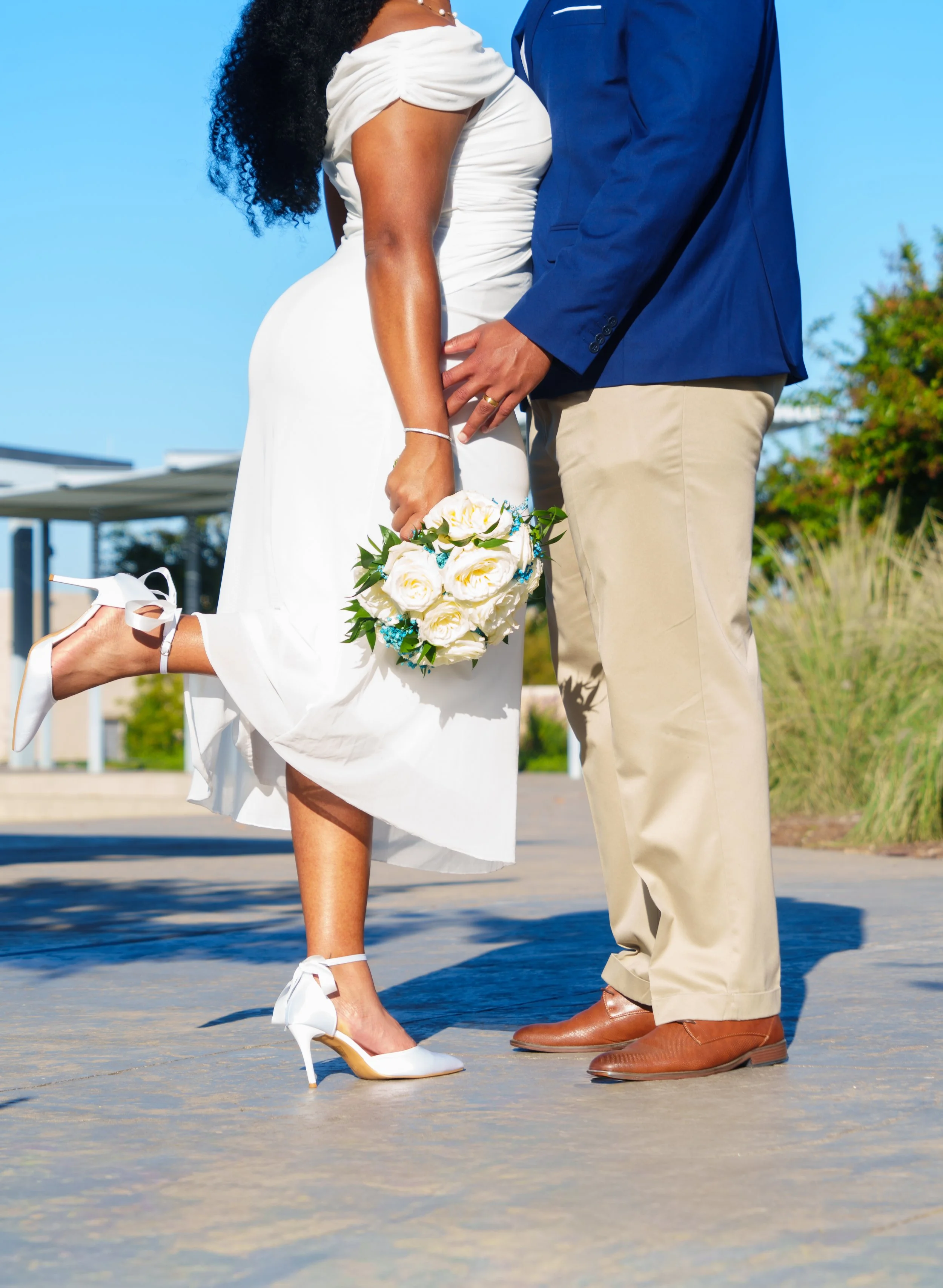 Close-up of a couple on their wedding day, with the woman wearing a white dress and holding a bouquet and the man dressed in a blue blazer and beige pants, standing outdoors under a sunny sky.
