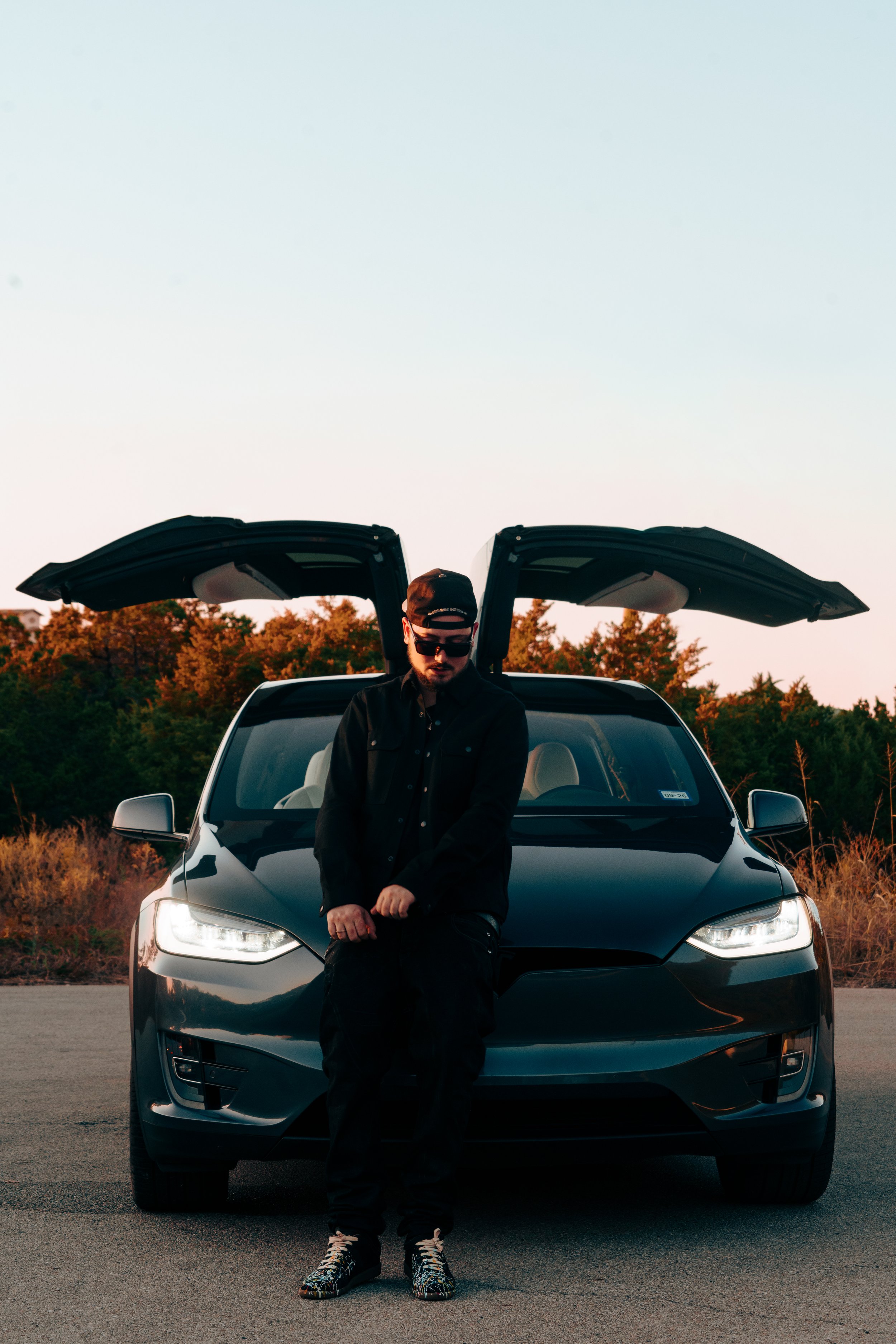 A young man dressed in black clothing and accessories, sitting on the front bumper of a black electric vehicle with gull-wing doors open in an outdoor setting during sunset.