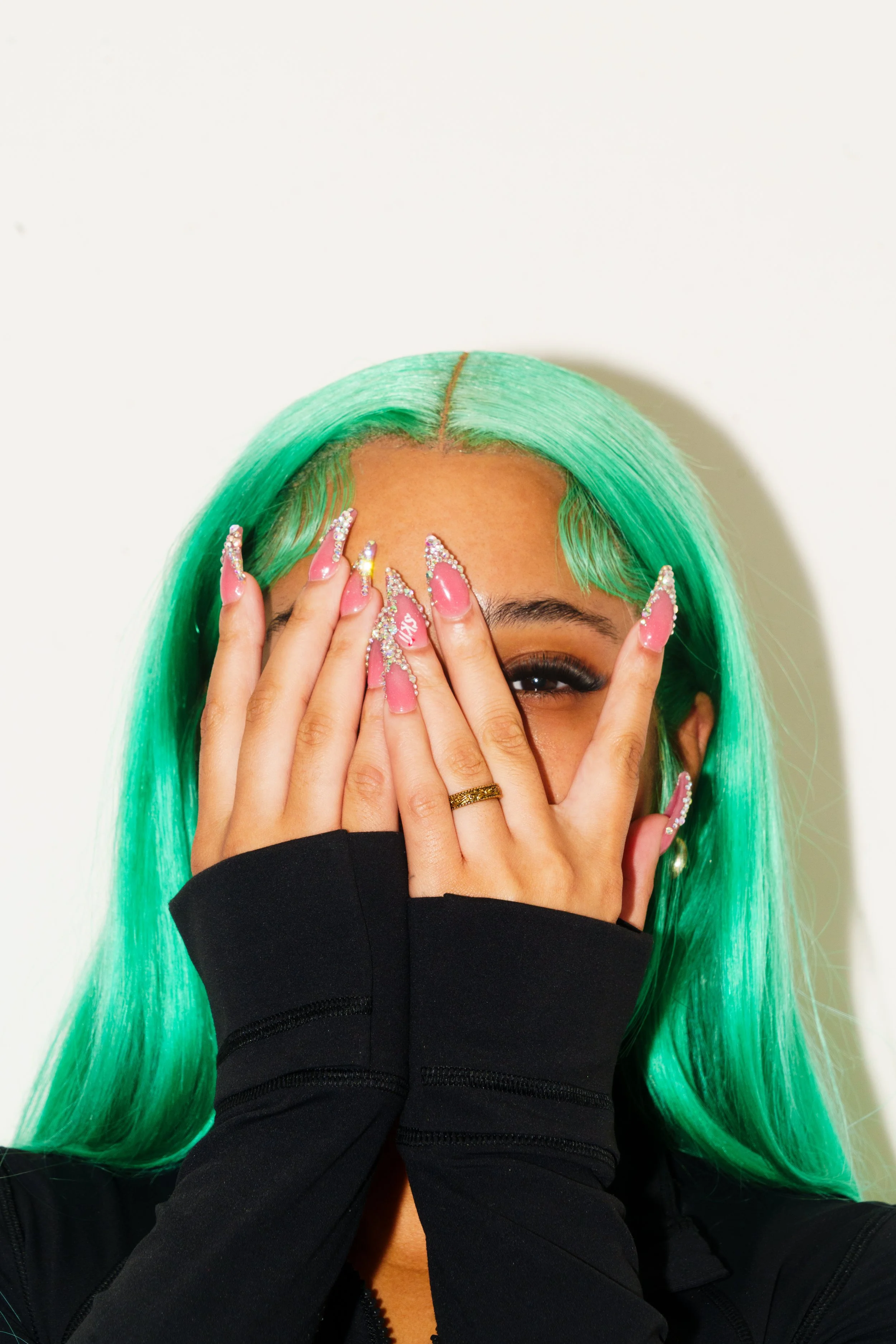 A person with neon green hair covering part of their face with their hands, which have long pink nails decorated with rhinestones. The person is wearing a black jacket and a gold ring.
