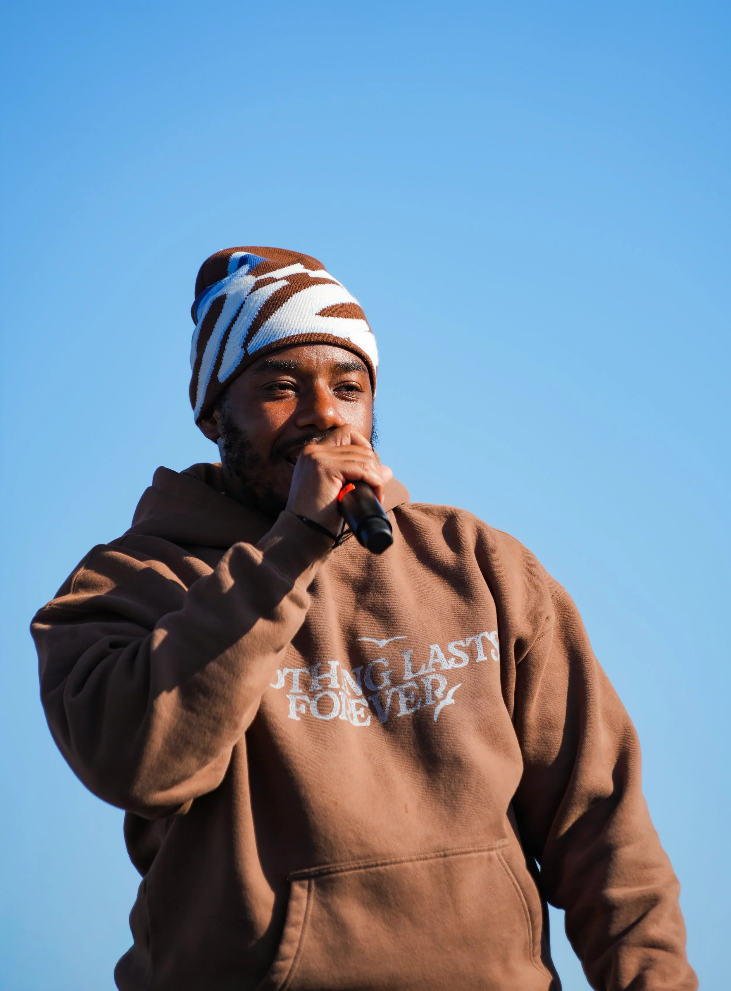A man wearing a brown hoodie and a multicolored beanie hat, holding a microphone to his mouth, against a clear blue sky.