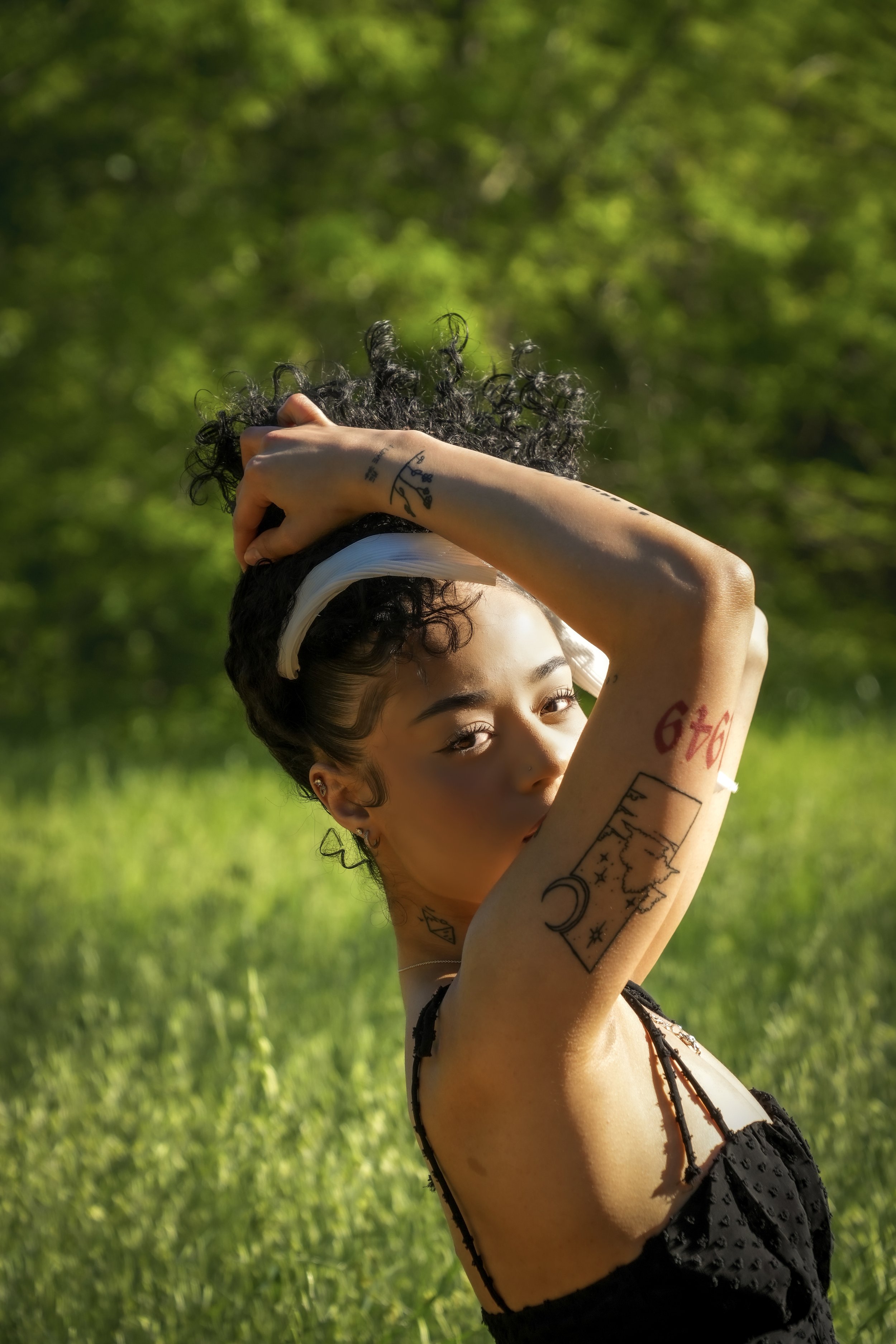 Young woman with tattoos on her arm, wearing a white headband and a black top, holding her curly hair with one hand, outdoors in a green setting.