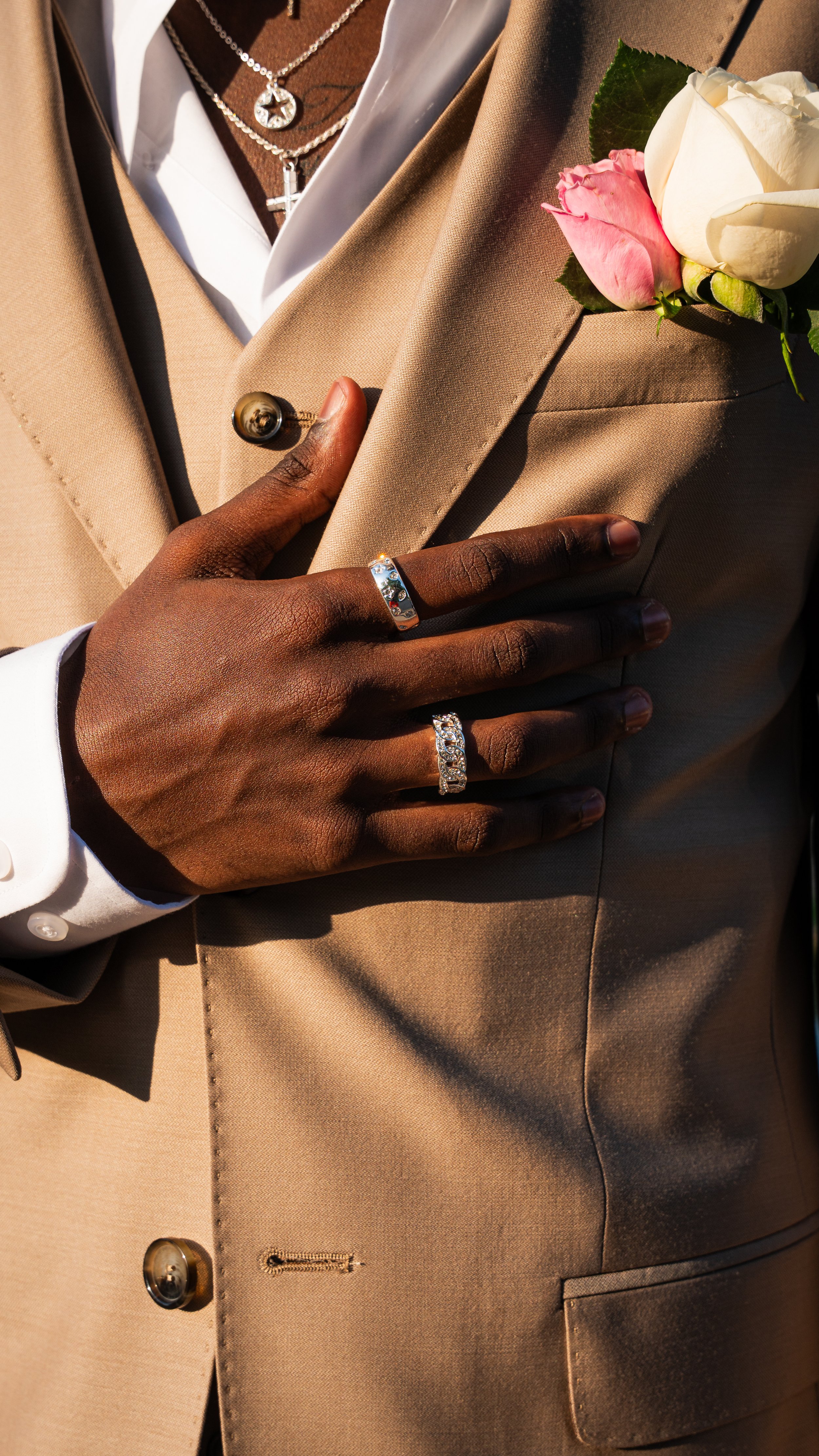 A person wearing a tan suit with a white shirt, four rings on their fingers, and a silver necklace with a cross and a star pendant. They are holding their chest with one hand, displaying a boutonniere with a pink and white rose.