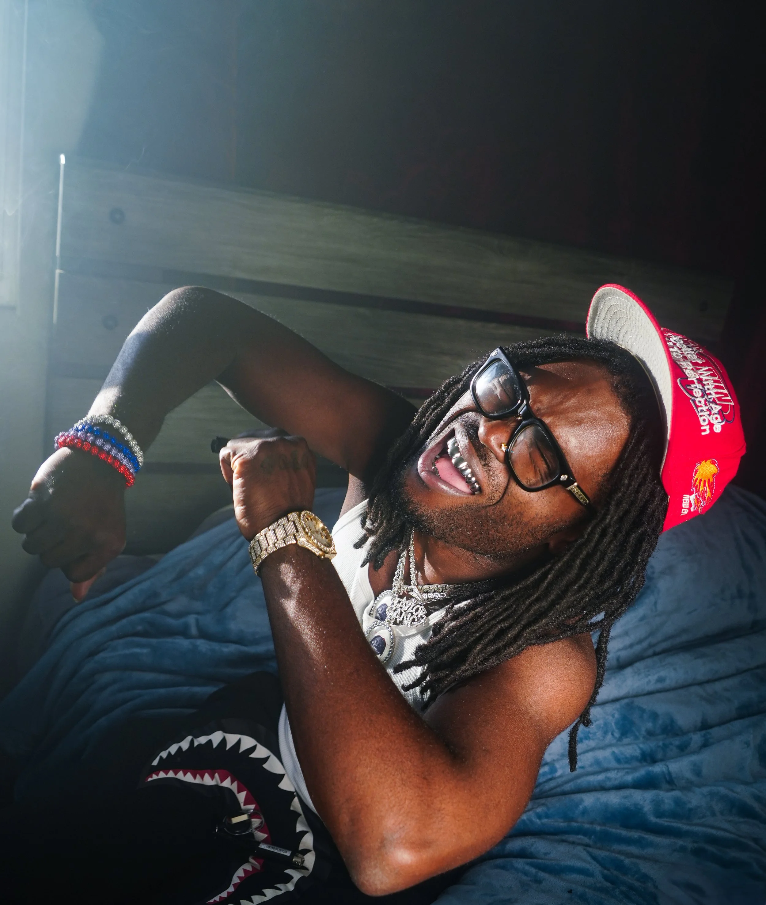 A man with long dreadlocks, glasses, and a red cap is lying on a bed, smiling, wearing gold jewelry and colorful bracelets.