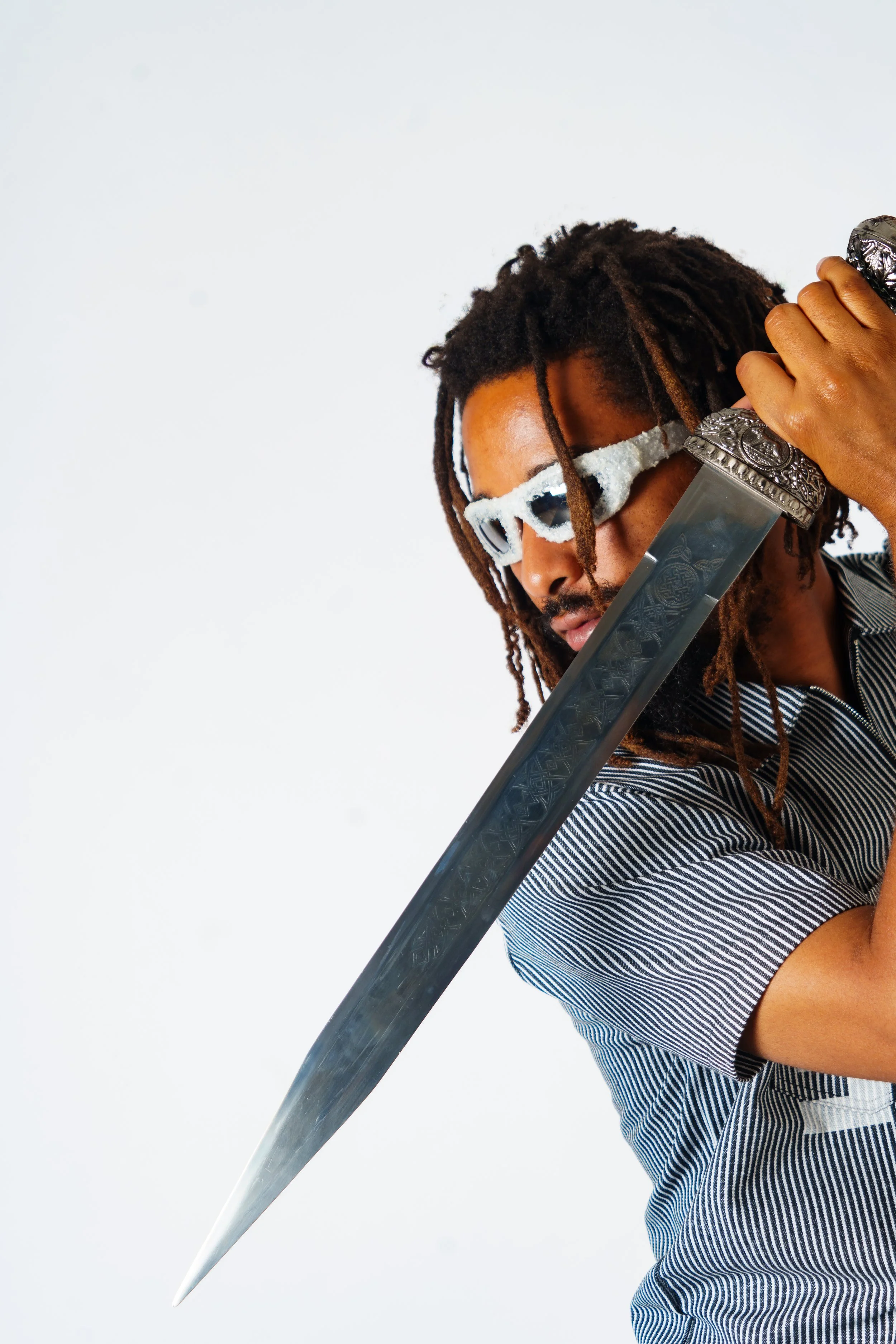A man with dreadlocks wearing sunglasses and a striped shirt holding a large sword.